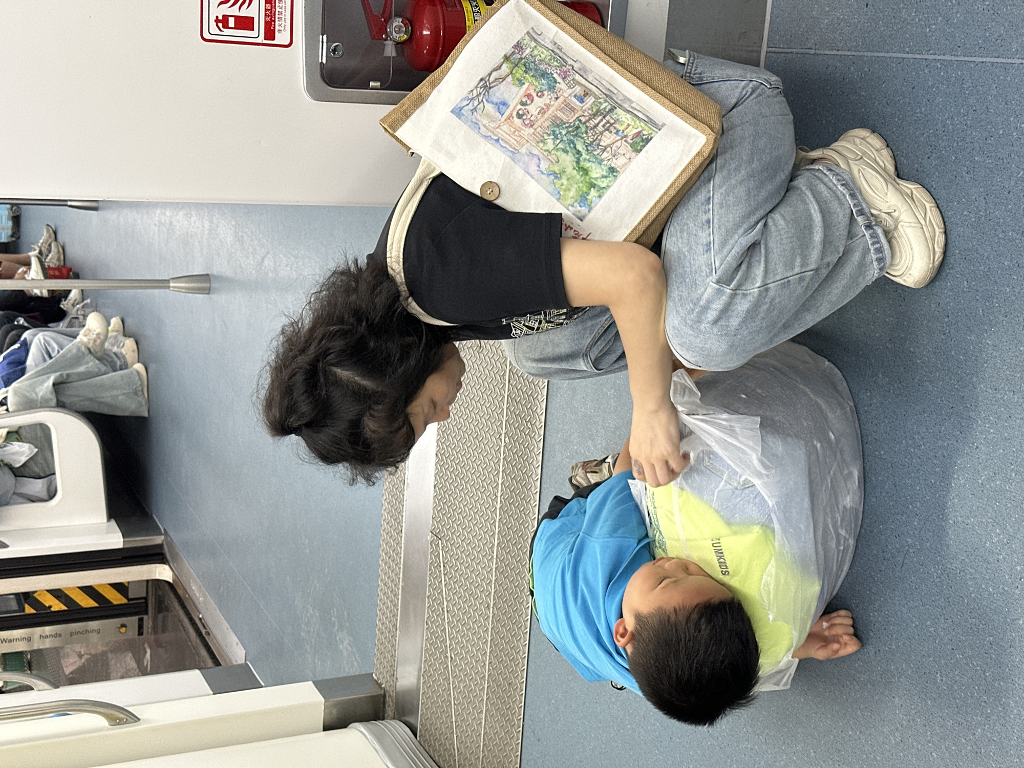 A medium, eye-level shot depicts an adult and a child indoors, possibly in a subway car. The adult, with dark curly hair, wears a black top and light-wash jeans. They are kneeling and looking down, appearing to be interacting with the child. A shoulder bag with a painting or drawing is slung over their shoulder.
The child, wearing a blue hoodie and a bright yellow t-shirt with the word "UMKIDS" visible, is sitting on the floor inside a clear plastic bag. The adult's hand is reaching into the bag towards the child.
The environment features a gray non-slip floor, and a metal staircase. Additional details include a fire extinguisher mounted on a wall, a sign with Chinese characters and safety warnings. Part of the subway car’s interior, including a black and yellow striped warning sign, is visible.