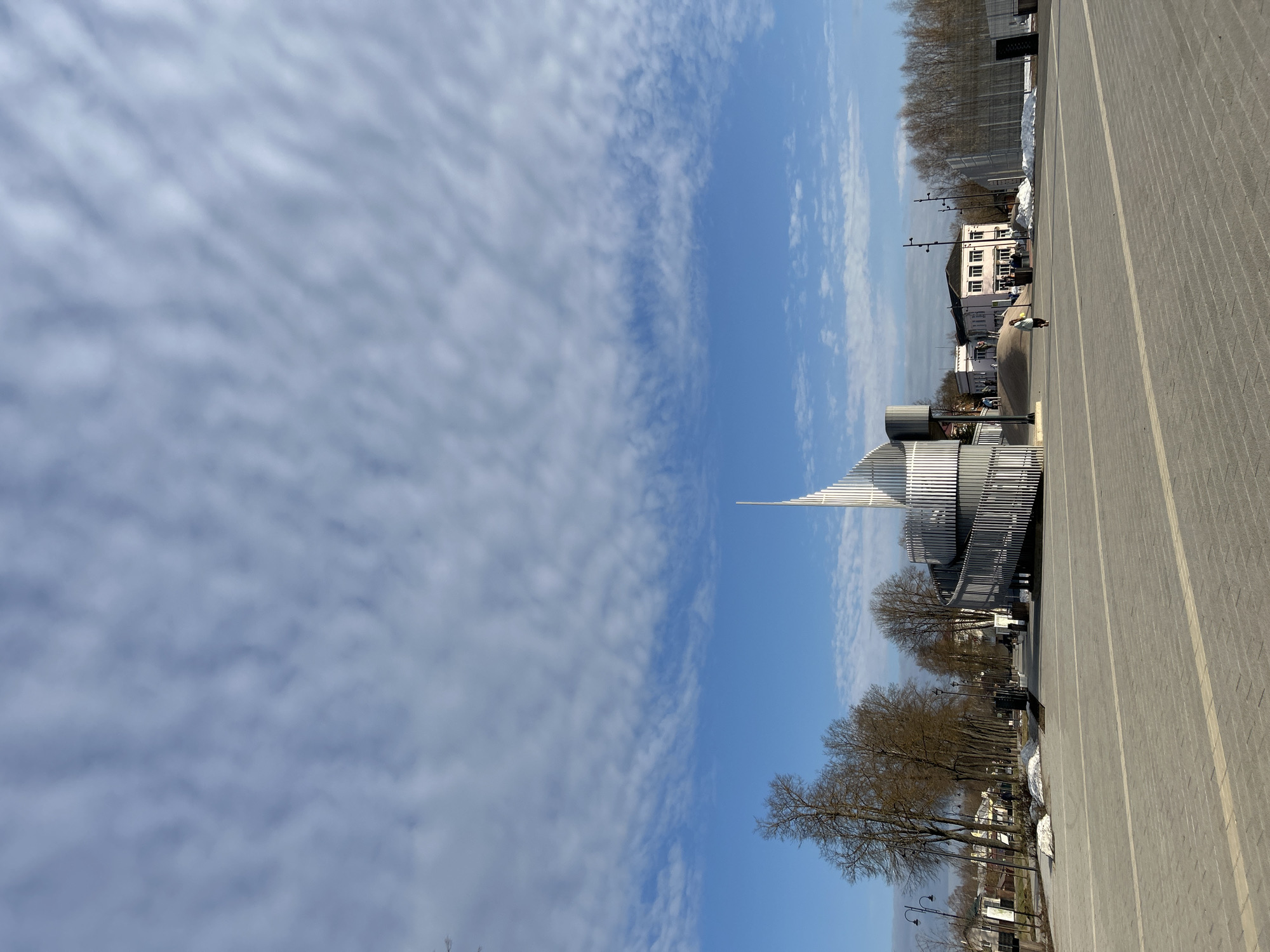 This image depicts a wide-angle view of the Memory Alley memorial complex in Klin, Russia, under a partly cloudy blue sky. The foreground consists of a large, light grey paved plaza composed of rectangular pavers, with two distinct lighter grey linear bands running horizontally across the lower third of the frame.
The central subject is a modern, metallic memorial structure, distinguished by a spiraling, ribbed ramp that ascends around a central, pointed spire. The structure is predominantly silver-grey. Several individuals are visible on or near this structure: two figures are seen ascending the spiral ramp, and a small group of approximately three to five individuals stands at its base, near an entrance point.
To the left of the plaza, a treelined avenue is visible, featuring bare deciduous trees with dark bark and several multi-armed streetlights. Patches of melting snow are present along the edge of the paved area. In the background, behind the trees, various buildings are partially visible.
To the right of the central memorial, a low-rise, light-pink building with multiple rectangular windows is situated on a slight incline. Several people are discernible near its entrance. Further to the right, a tall metal fence or barrier is present, with more bare trees behind it and additional patches of snow.
One individual, dressed in light outerwear, is walking alone across the open paved plaza from the left foreground towards the right midground, heading in the general direction of the memorial structure. The sky overhead features a blend of deep blue and scattered, wavy white clouds, which become denser and more uniform towards the top of the frame. The lighting indicates daytime, with shadows suggesting the sun is to the left of the camera's perspective. The presence of bare trees and remnants of snow suggests a late winter or early spring season.
