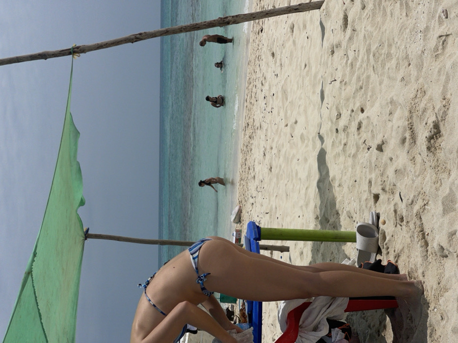 The image captures a beach scene in Zanzibar, Tanzania, under an overcast sky. In the foreground, a person, likely female, with light skin, is seen from the waist down, bent over and facing away from the viewer. They are wearing a blue and white striped bikini bottom and the top is partially visible. This individual appears to be interacting with items on or near a small blue and green plastic table. Beneath the table, a red container is partially visible, draped with white fabric. A single white slide sandal and a black sandal rest on the light-colored sand.
A makeshift green tarp shade structure is supported by crude wooden poles, one of which anchors the right side of the frame. The sandy beach extends towards the clear, turquoise ocean in the midground, where four individuals are present in the shallow water. One person, with dark hair, walks left into deeper water. Another person, also with dark hair, stands facing right. A third person is partially submerged, with only their head visible above the water. The fourth individual, likely male, stands facing left, looking down into the water. The ocean horizon is visible under a light grey sky.