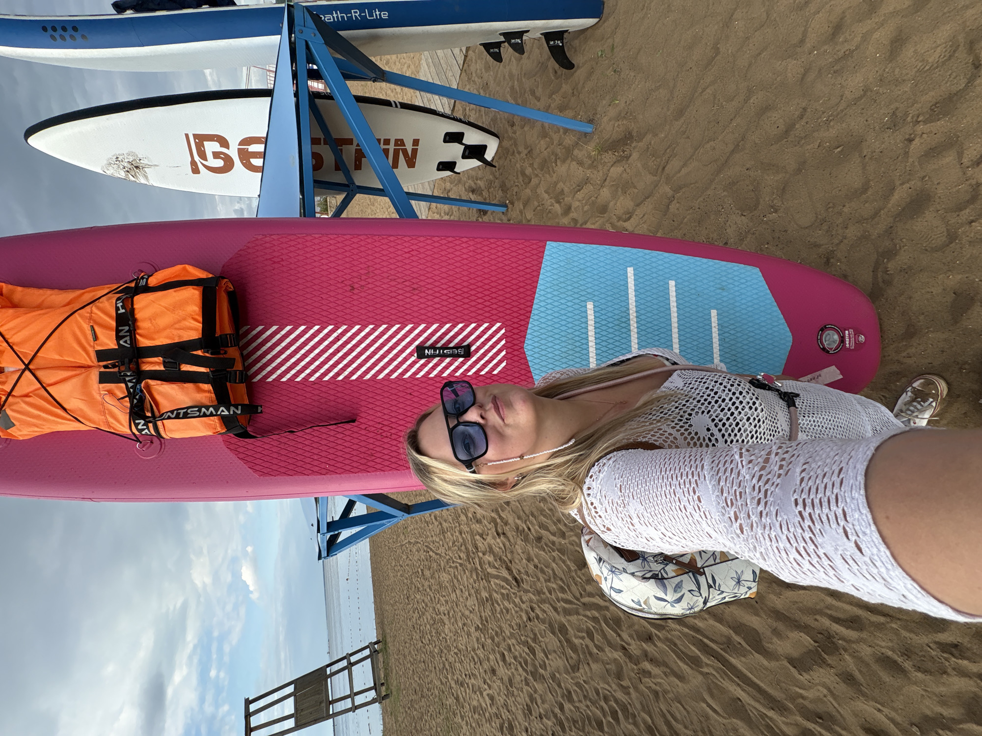 A blonde woman wearing sunglasses, a white crochet cover-up, and a patterned backpack stands on a sandy beach in Chaikovsky, Russia, next to a pink paddleboard with an orange life vest attached. The paddleboard is positioned vertically, and the woman is taking a selfie. In the background, there are other paddleboards on a blue metal rack, a wooden lifeguard tower, and a body of water under a cloudy sky. The sand has visible footprints and ripples.