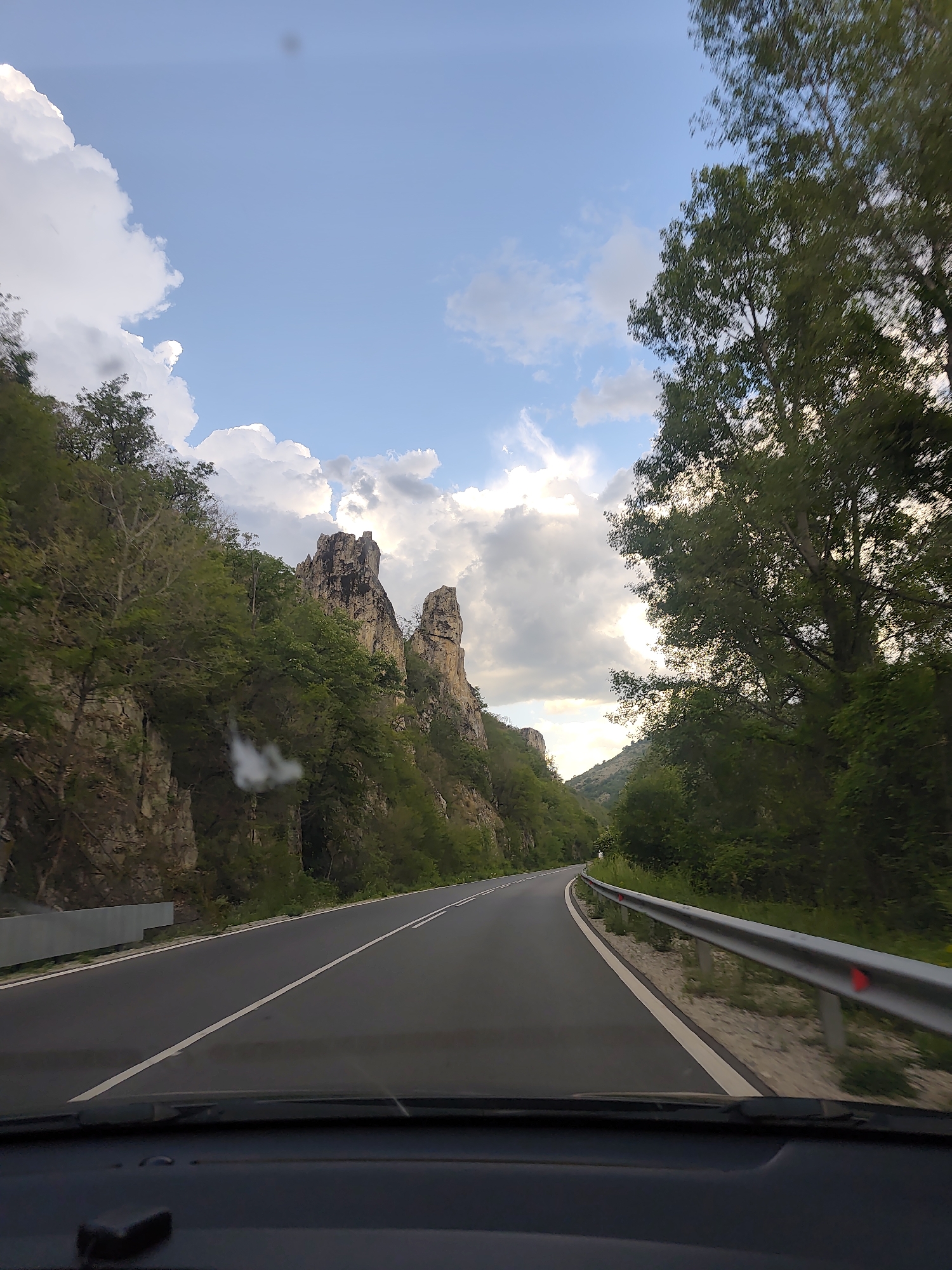 The image depicts a view from inside a vehicle driving on a paved road. The road curves slightly to the right, with a metal guardrail visible on the right side. A rock formation is visible in the background. The sky is visible above, featuring clouds. The image was taken on the road to Gospodintsi, Bulgaria.