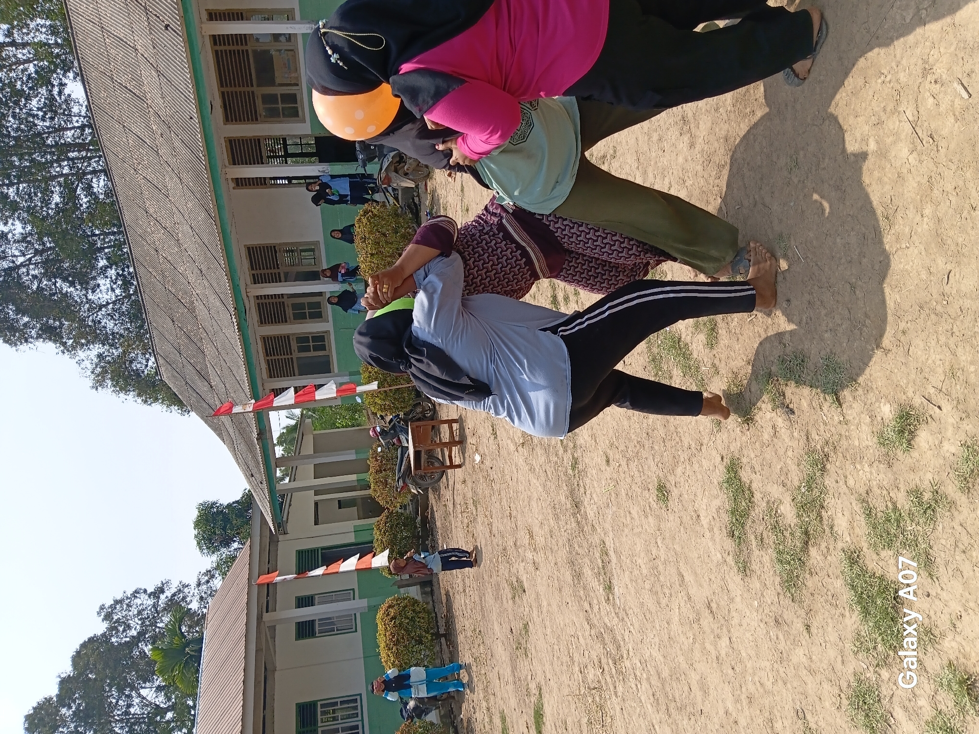 The image shows an outdoor scene in Margo Rukun, Indonesia, where a group of women are engaged in a game. In the foreground, three women are actively participating in what appears to be a tug-of-war or a similar strength-based activity, holding onto each other and bracing themselves on the sandy ground. They are all wearing headscarves. One woman is wearing a light blue shirt and black pants with white stripes, another is in a patterned dress and green pants, and a third is in a bright pink shirt and black pants. Two balloons, one orange and one green, are visible near the women. In the middle ground, two younger girls in hijabs are observing the activity. Further back, more people are visible near a building with a corrugated metal roof and white walls, decorated with Indonesian flags. The environment is a dirt ground with some patches of grass and dry vegetation. Two motorcycles are parked near the building, and a small wooden table is positioned on the ground. The scene is under a bright, clear sky. The watermark "Galaxy A07" is present in the lower left corner.