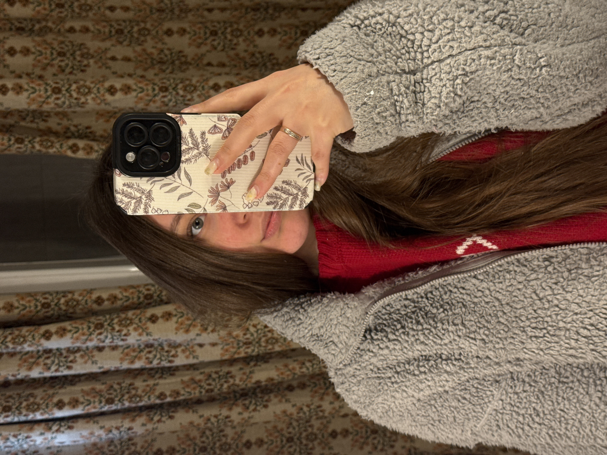 The image captures a self-portrait of a person indoors, holding a smartphone to partially obscure their face. The subject is a woman or young adult with long, dark brown hair. Her left eye, appearing light blue or grey, is visible, along with parts of her nose and pink lips. She is dressed in a grey, textured, sherpa-style jacket with a visible zipper, worn over a red sweater that shows a small white pattern near the neckline.
In her right hand, she holds a smartphone, identifiable by its triple-camera setup, enclosed in a cream or beige-colored case. The phone case features a delicate botanical pattern with illustrations of leaves, small flowers, and possibly butterflies or moths. A gold-colored ring, adorned with small, sparkling stones, is worn on the ring finger of her right hand. Her fingernails are a natural, light color.
The background consists of pleated curtains or fabric, displaying a repeating intricate pattern in earthy tones of brown, orange, and cream. The scene suggests a casual, indoor setting. There is no visible text in the image. The time of day is not discernible, but the jacket implies cooler temperatures.