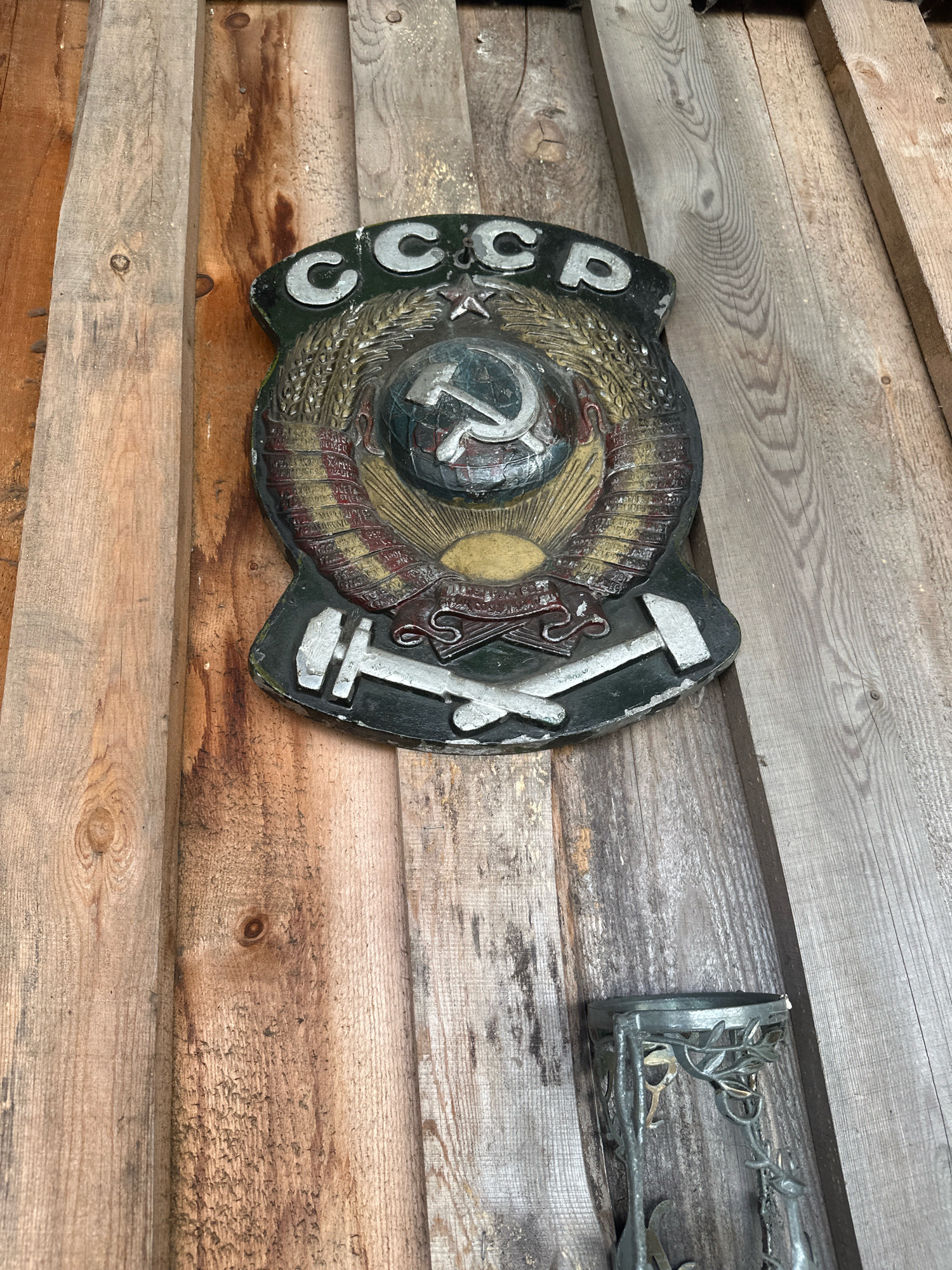 The image shows a close-up, high-angle shot of a weathered Soviet crest mounted on a wooden wall. The crest, featuring the hammer and sickle and the Cyrillic letters "СССР" (USSR), is the central subject. The wooden wall is constructed from rough-hewn planks, some with visible knots and grain. The overall color palette is muted, dominated by the browns of the wood and the faded colors of the crest. In the lower right corner, a portion of a decorative metal object, possibly a candle holder or lantern, is visible. The scene appears to be outdoors, given the natural lighting and the weathered appearance of the materials. There is no indication of Kargat, Russia, or any specific location.