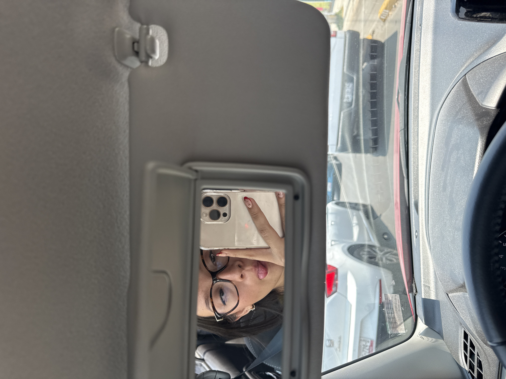 A woman is seen in a rearview mirror, reflected in a car's sun visor. She is wearing glasses and holding up a smartphone, presumably taking a selfie. The car appears to be stationary, possibly in traffic, as other vehicles are visible through the windshield. The location is in San Bernardino Tlaxcalancingo, Mexico. The dashboard, steering wheel, and interior details of the car are visible.
