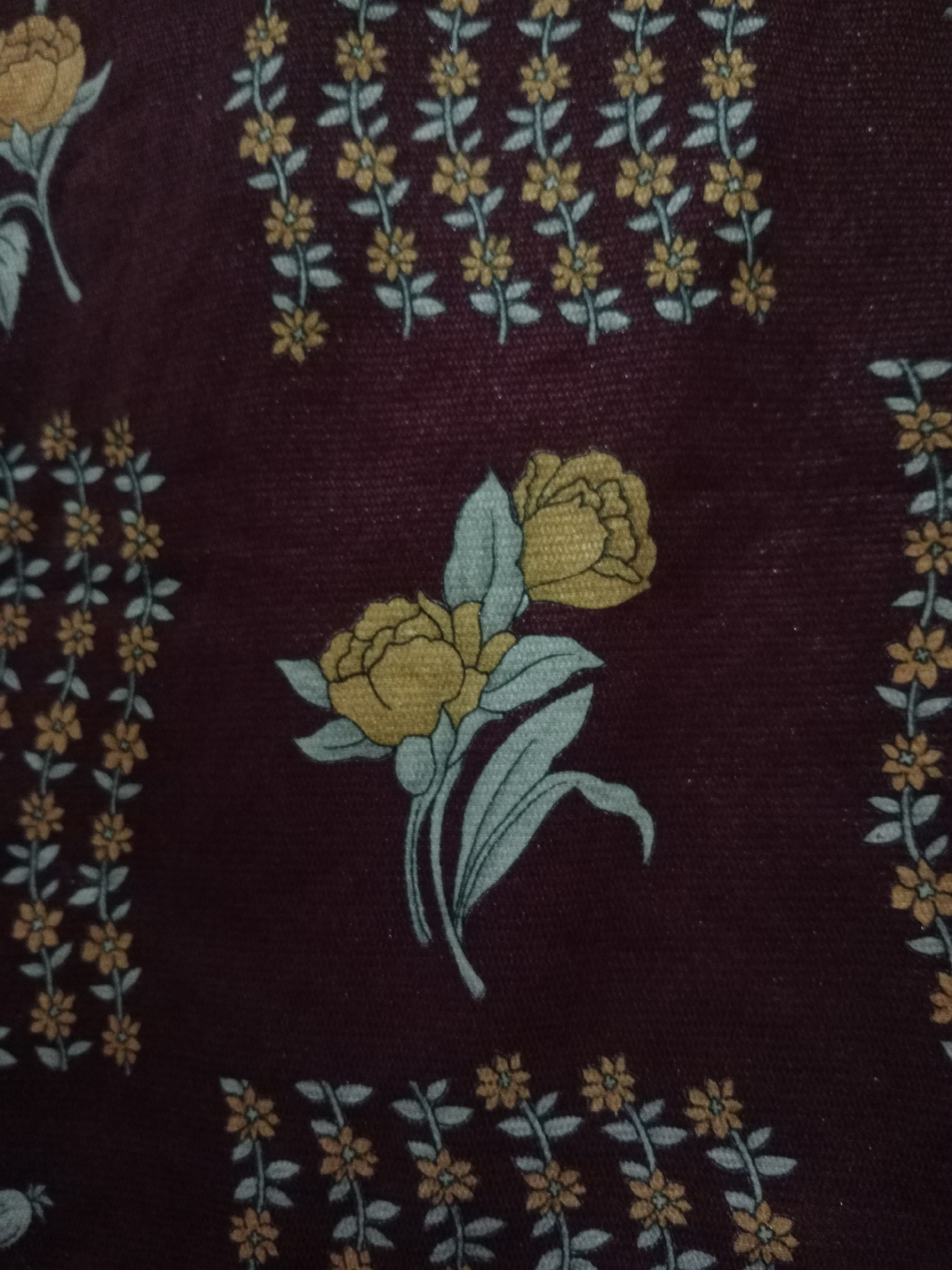 The image shows a close-up of a dark maroon fabric with a floral pattern. The pattern consists of rows of small, light yellow flowers with light teal stems and leaves. There are also larger floral motifs, including a bunch of two light yellow roses with teal leaves and stems. The fabric appears to have a slight texture, possibly from the weave or a nap. The lighting is soft and diffused, with no harsh shadows. The overall impression is of a decorative textile. No information about the location in the city is available from the image.