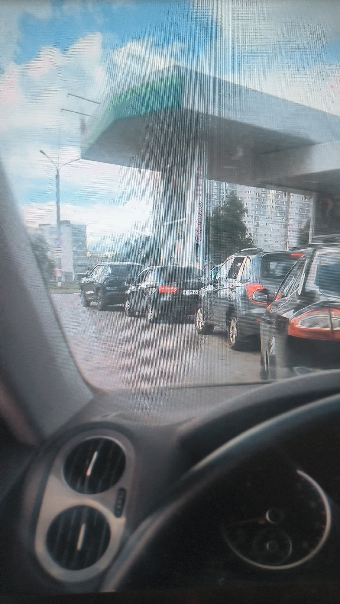 The image depicts a queue of cars at a gas station in the city of Kovrov, Russia. The perspective is from inside a vehicle, looking forward through the windshield. A modern gas station canopy with green accents is visible overhead. Several black cars are lined up on the asphalt, with one grey SUV in the middle of the queue. The background shows apartment buildings and a cloudy blue sky. The interior of the vehicle shows part of the steering wheel and dashboard, including air vents. The image quality is somewhat degraded with vertical lines, suggesting it might be a photo of a screen or taken with a low-quality camera.