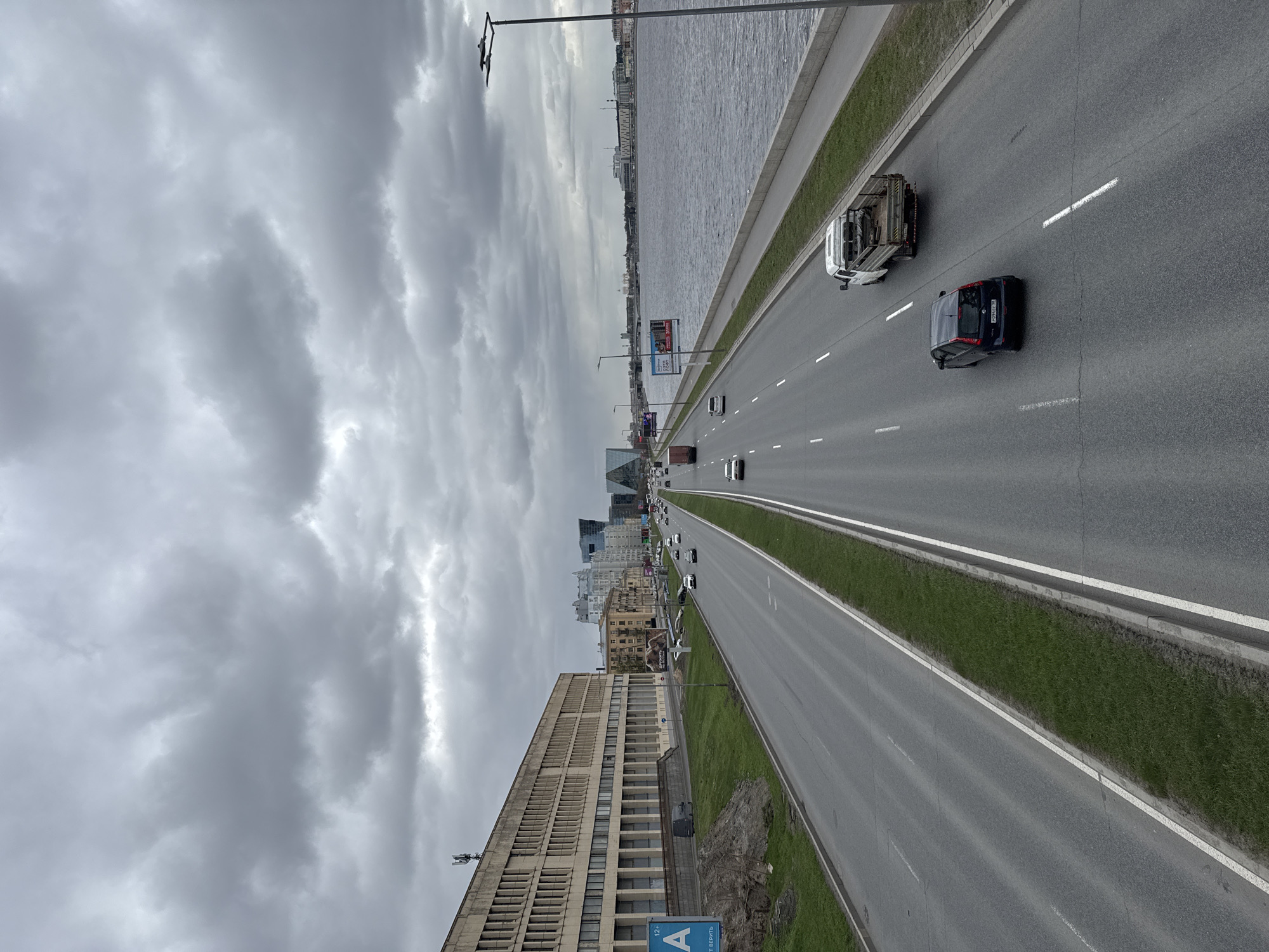 This aerial shot depicts a multi-lane highway bisecting Saint Petersburg, Russia, under a cloudy sky. The Neva River parallels the highway, with buildings visible in the distance. Traffic is light, with a mix of cars and trucks present. On one side of the highway is a large, modern building with visible windows, suggesting an office or residential complex. A grassy median divides the traffic lanes. A sign with the letter "A" in blue is visible near the road.