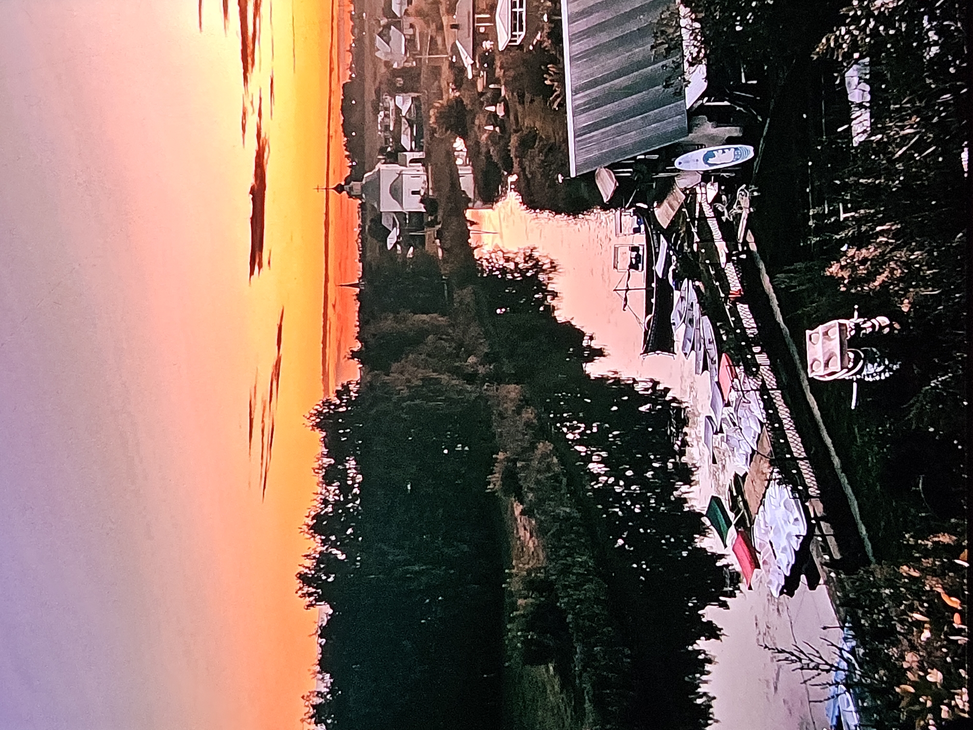The image depicts a serene sunset scene by a river, likely in or near Vladimir, Russia, given the presence of a white domed church and other traditional Russian architecture. The sky transitions from a soft violet at the top to vibrant orange and yellow near the horizon, with wispy clouds tinged with red. On the left bank of the river, a dense line of dark trees stands silhouetted against the colorful sky. The river itself, calm and reflective, mirrors the trees and the warm hues of the sunset.
Along the riverbank on the right, a small village is visible, characterized by several houses with pitched roofs and a prominent white church with a golden dome and cross. The architecture suggests a historical Russian setting. Moored along the river's edge are numerous small boats, including paddle boats and some larger, darker vessels. Some of these boats are white, while others are red or green. A fence runs along the embankment where the boats are docked. In the foreground, a weathered, dark-colored building with a corrugated metal roof is partially visible, along with a paddleboard leaning against it. The overall atmosphere is peaceful and idyllic, capturing the beauty of a rural Russian evening.