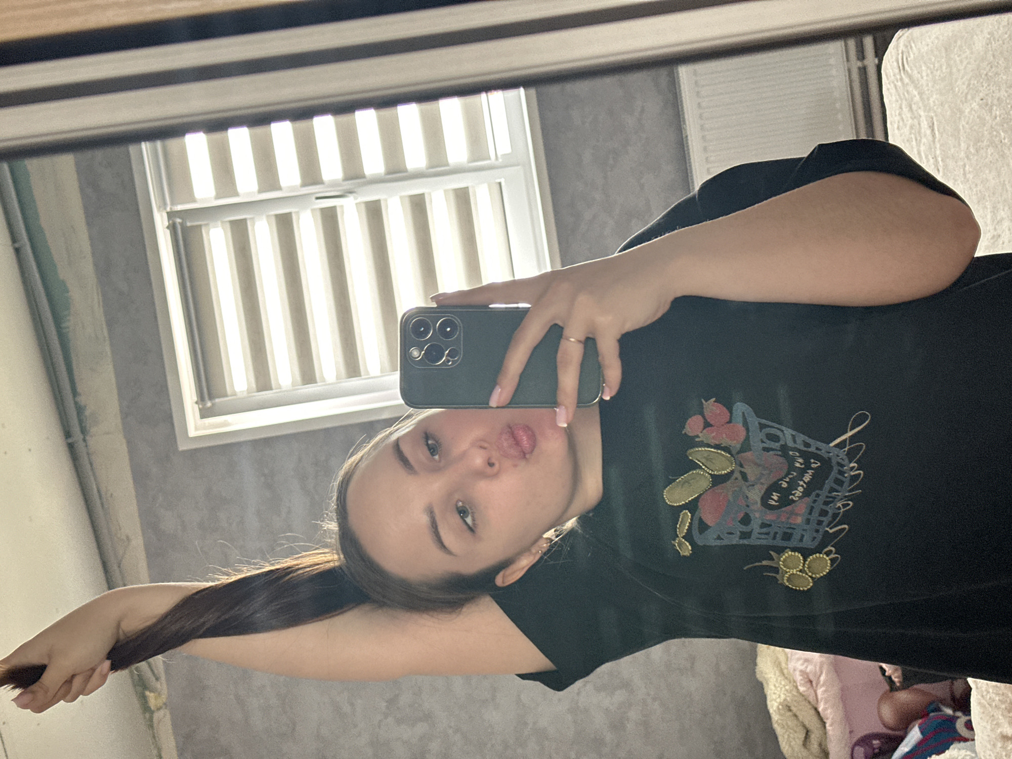An indoor mirror selfie captures a young woman with long, dark hair, wearing a black t-shirt. The image is rotated 90 degrees clockwise. She holds a dark green or black smartphone with a triple-lens camera setup, visible in the mirror reflection as she takes the photo. Her right hand, adorned with a simple ring, holds the phone. Her nails appear manicured with a light polish.
The woman looks directly into the camera, pouting her lips in a playful expression. Her eyes are light-colored, and she has visible dark eyebrows.
Her black t-shirt features an embroidered or printed design on the front, depicting a basket containing stylized fruit (berries, possibly avocados or similar shapes) and golden embellishments. Within the basket, there is stylized text that is partially legible; it appears to contain words like "MOMENT" or "LOVE ME".
The background shows an indoor residential setting, likely a room with light grey, possibly textured, walls. Behind the woman, a window with horizontal blinds or slats allows natural daylight to enter, indicating it is daytime. To the right of the window, a radiator is partially visible. In the bottom right corner, a glimpse of light-colored bedding or blankets suggests a bed or sofa.