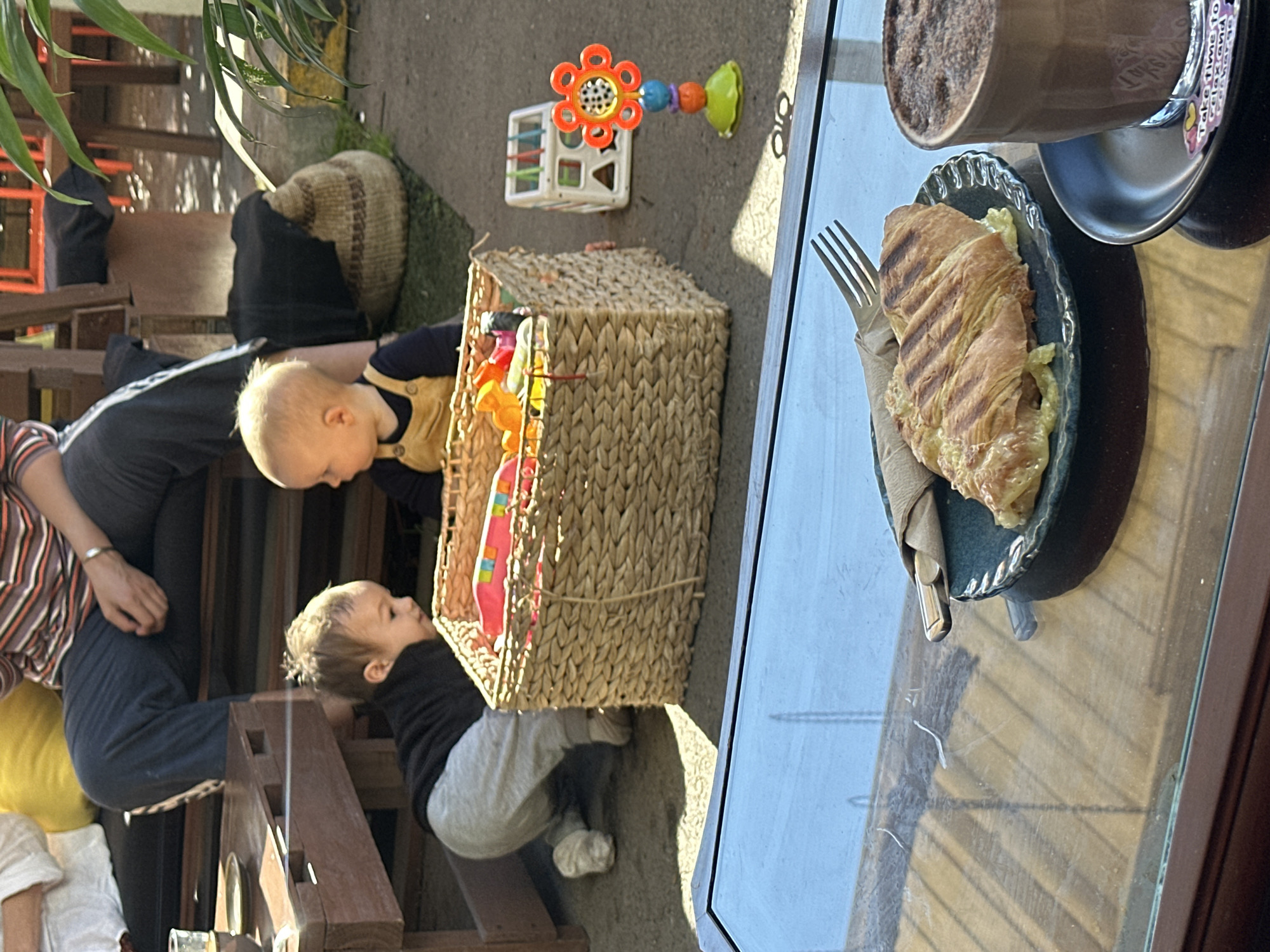 The image shows a sunny outdoor seating area at a cafe in Bellingen, Australia. In the foreground, a glass table reflects the environment. On the table rests a dark blue plate holding a grilled pastry and a fork, next to a glass of chocolate drink. Behind the table, two babies are playing near a woven basket filled with toys. One baby, wearing a black top and grey pants, is on its hands and knees, looking towards the basket. The other baby, wearing a yellow outfit, sits facing the first baby. In the background, an adult is seated, their legs visible, with a large woven basket and some outdoor furniture. The ground appears to be asphalt or concrete, with some grass and plants visible. The overall impression is a relaxed, casual atmosphere, likely a cafe or a backyard.