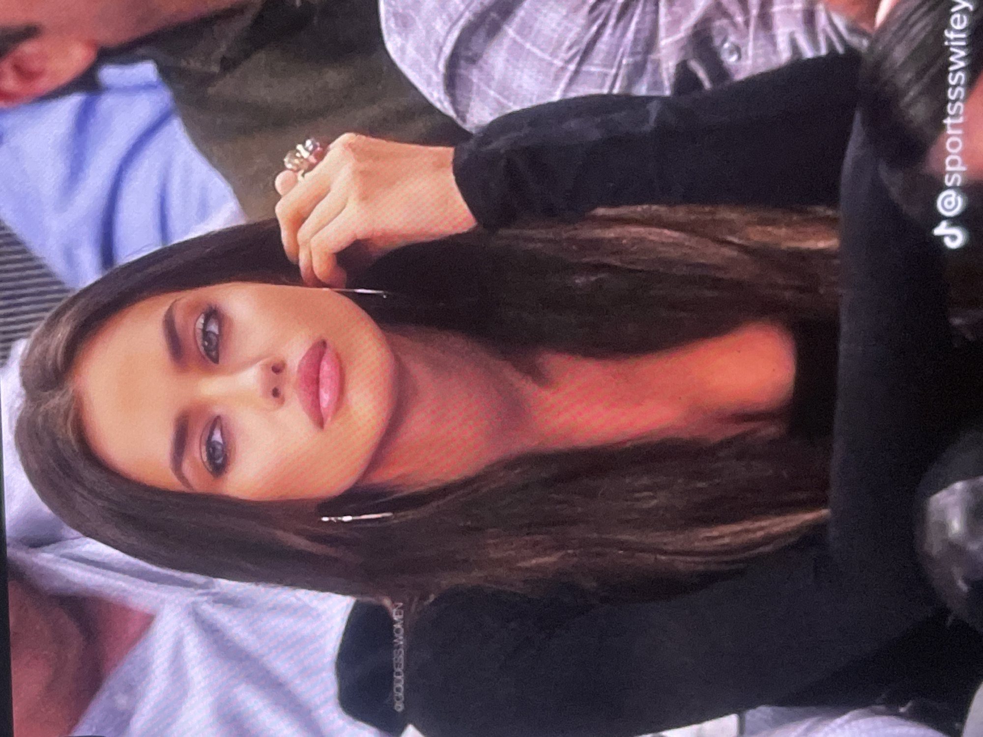 The image displays a young woman looking directly at the viewer, with the image itself rotated 90 degrees counter-clockwise. She has long, dark brown hair, prominent eyebrows, and is wearing makeup, including pink lipstick and eye makeup. She wears large hoop earrings and a distinctive ring on the ring finger of her right hand, which is positioned near her head. Her attire consists of a simple black long-sleeved top.
In the blurred background, parts of other individuals and their clothing are visible, suggesting an indoor public setting, possibly an event or a gathering. One person behind her appears to be wearing a light-colored, possibly plaid or checked, button-up shirt. The overall image quality indicates it might be a screenshot or a photo taken of a screen, showing some pixelation.
The woman maintains a composed and confident expression.
Visible text includes a TikTok logo accompanied by the username "@sportssswifey" vertically oriented on the right side of the image. Another vertical text, "@GODDESS.WOMEN", is visible on the bottom left. The location context provided for this media file is Poltava, Ukraine, though no specific visual elements in the image directly indicate this location.