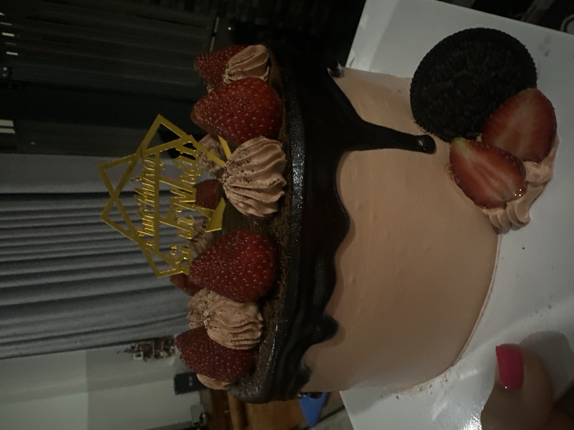 A cake decorated with strawberries, chocolate, and an Oreo cookie sits on a white surface. A gold cake topper is also present. The background shows a dimly lit room.