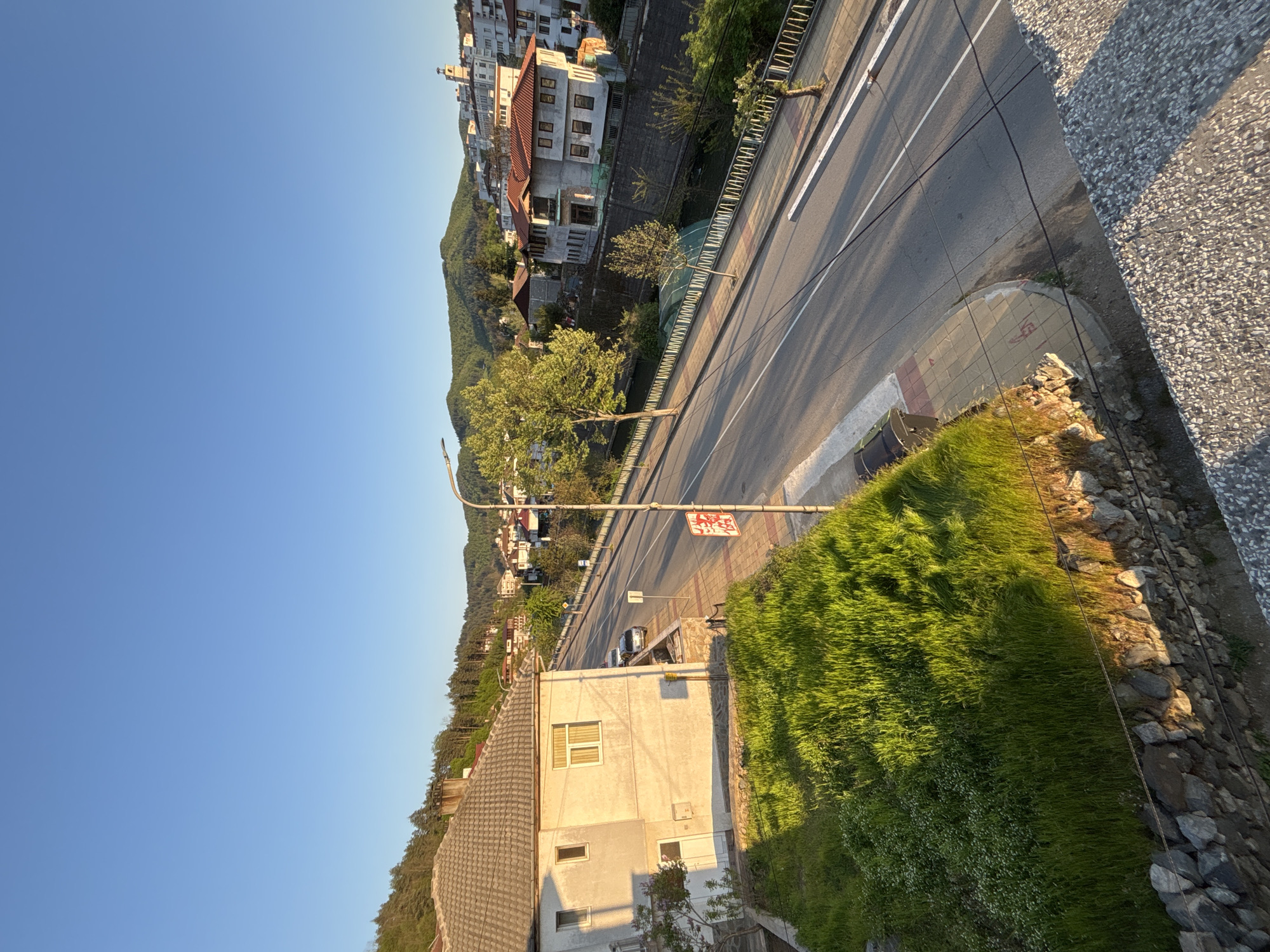 The image depicts a street scene in Zlatograd, Bulgaria, from an elevated perspective. A road runs through the scene, bordered by a metal railing on one side and a patch of grass and a building on the other. The building is light-colored with a gray roof. The road has painted lines. Beyond the road is a hillside with residential buildings. A traffic sign is visible near the road. The sky is clear and blue. The light suggests it might be early morning or late afternoon.