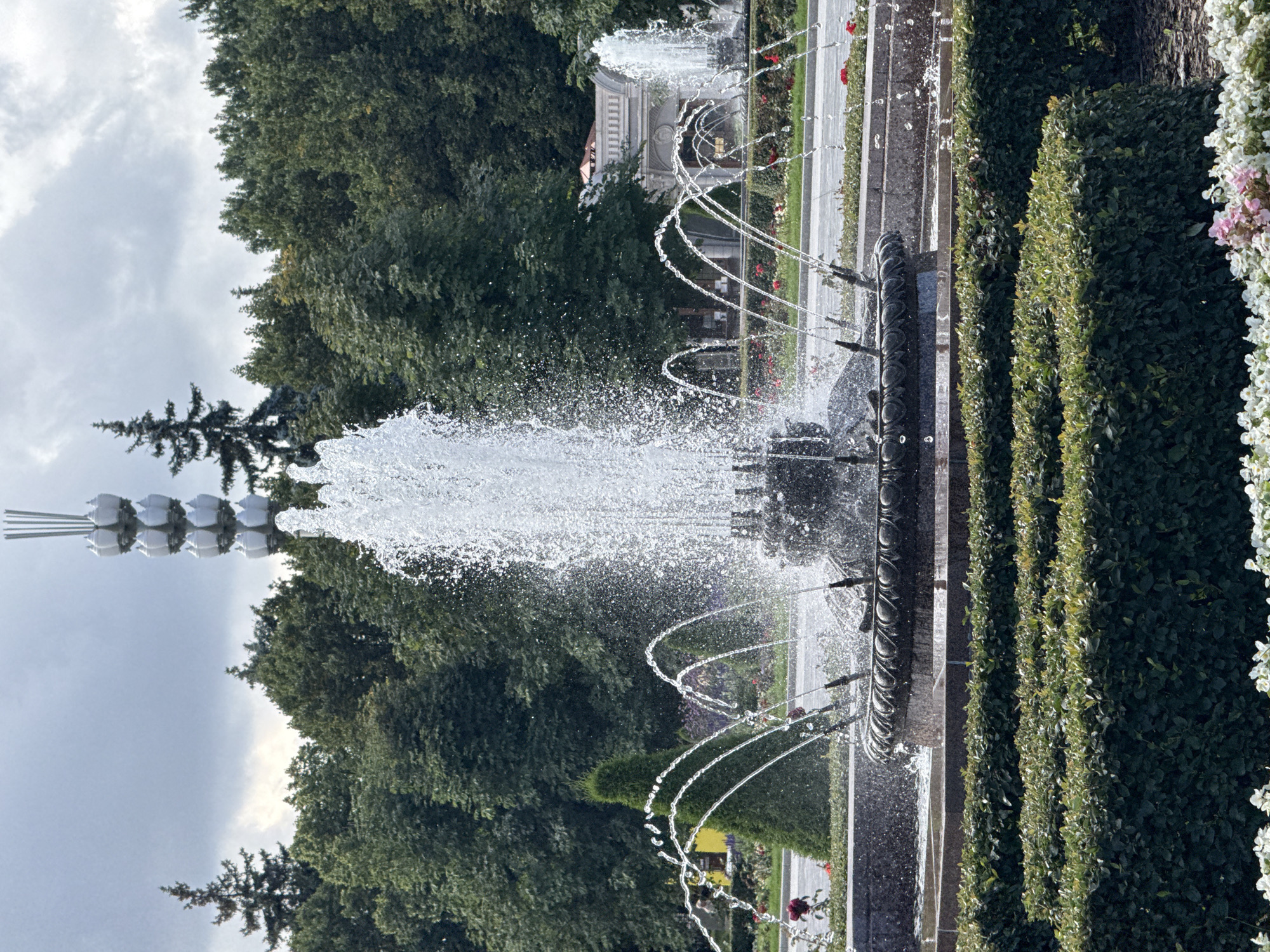 The image captures a view of the Central Moscow's architectural monument and fountain. The fountain, the main subject, is a large, multi-tiered structure with water spraying upwards and outwards in a dramatic display. It is surrounded by manicured greenery, including hedges and ornamental bushes, as well as some flowers in the foreground. In the background, a tall, slender tower with multiple lamp-like fixtures rises above a dense line of trees. The sky is overcast with clouds. The overall scene suggests a public park or square in a city setting, likely a place of leisure or historical significance.