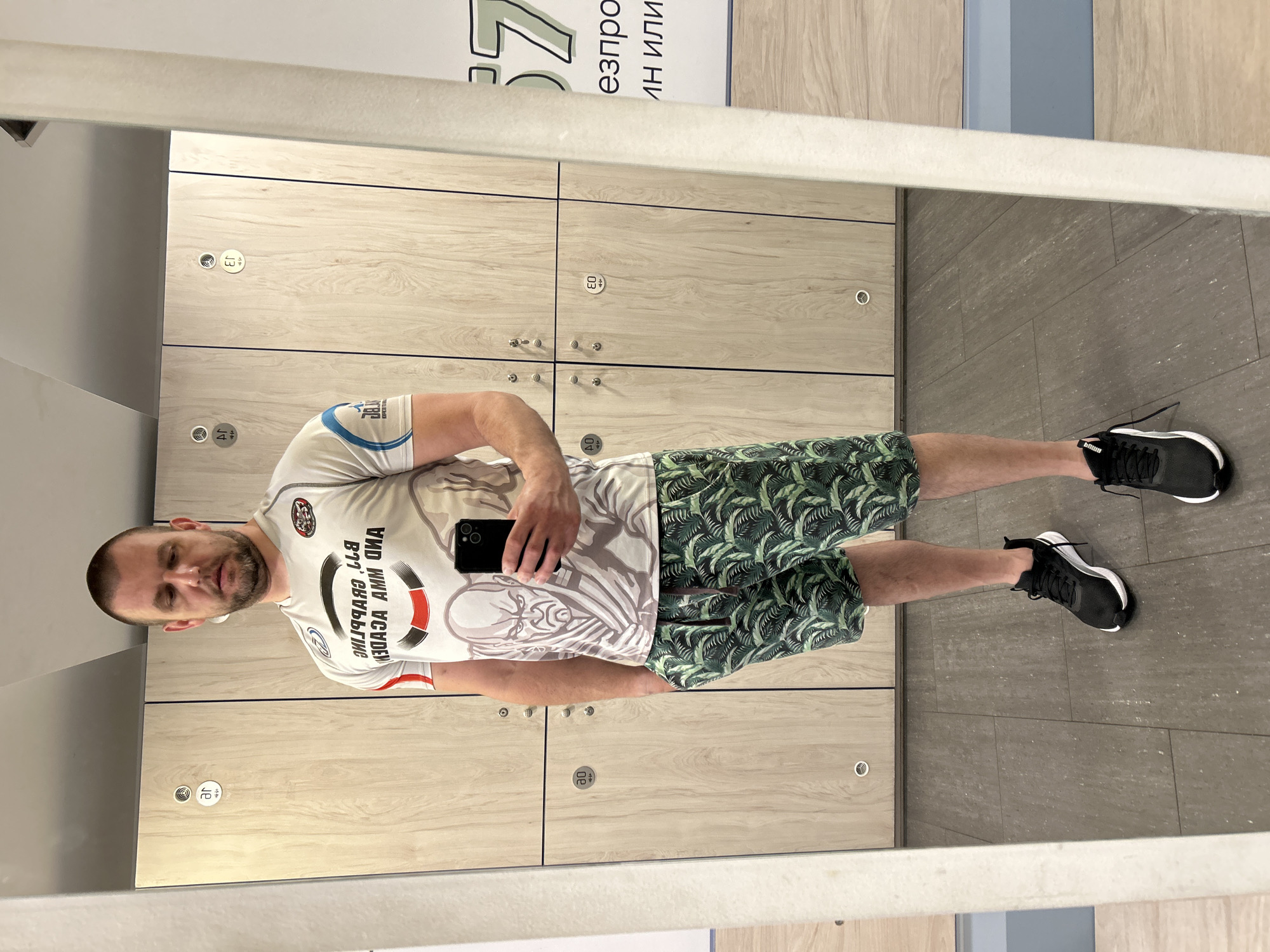A man is captured in a mirror selfie, standing in what appears to be a locker room or changing facility. He is holding a black smartphone in his right hand, taking the photograph. He wears a light-colored, short-sleeved t-shirt with graphics and reversed text, which appears to read "GRAPPLING" and "MMA ACADEMY". His lower attire consists of dark green shorts with a distinctive tropical leaf pattern, and he has black athletic shoes with white soles.
The setting features rows of light-colored wooden lockers with circular locks and numbers visible, including "03", "04", "J3", "J4", "20", and "40". The floor is tiled with rectangular, dark grey tiles. In the background, above the lockers, a sign with reversed Cyrillic text is partially visible, showing characters like "езпро", "ИШИ НИ", and the number "37". The man has a short beard and dark hair, and his expression is neutral, focused on the phone. The scene is brightly lit by indoor artificial lighting. The location is likely a gym or sports complex in Sofia, Bulgaria.