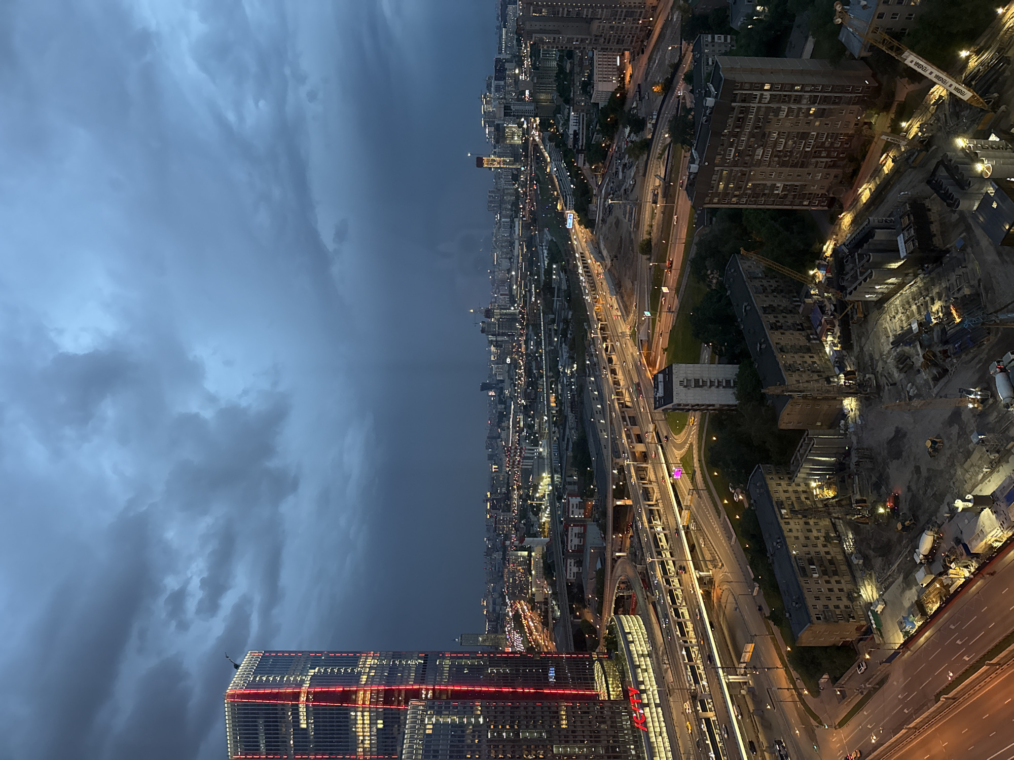 The image shows a high-angle view of a cityscape at dusk or night, likely in Khimki, Russia. The sky is overcast with dark, heavy clouds. In the foreground, a modern skyscraper with red horizontal bands is partially visible. Streets and highways below are illuminated with car headlights and streetlights. A dense urban area stretches into the distance. Construction sites with cranes and heavy equipment are visible. Several large buildings and residential areas are interspersed with highways and roads.