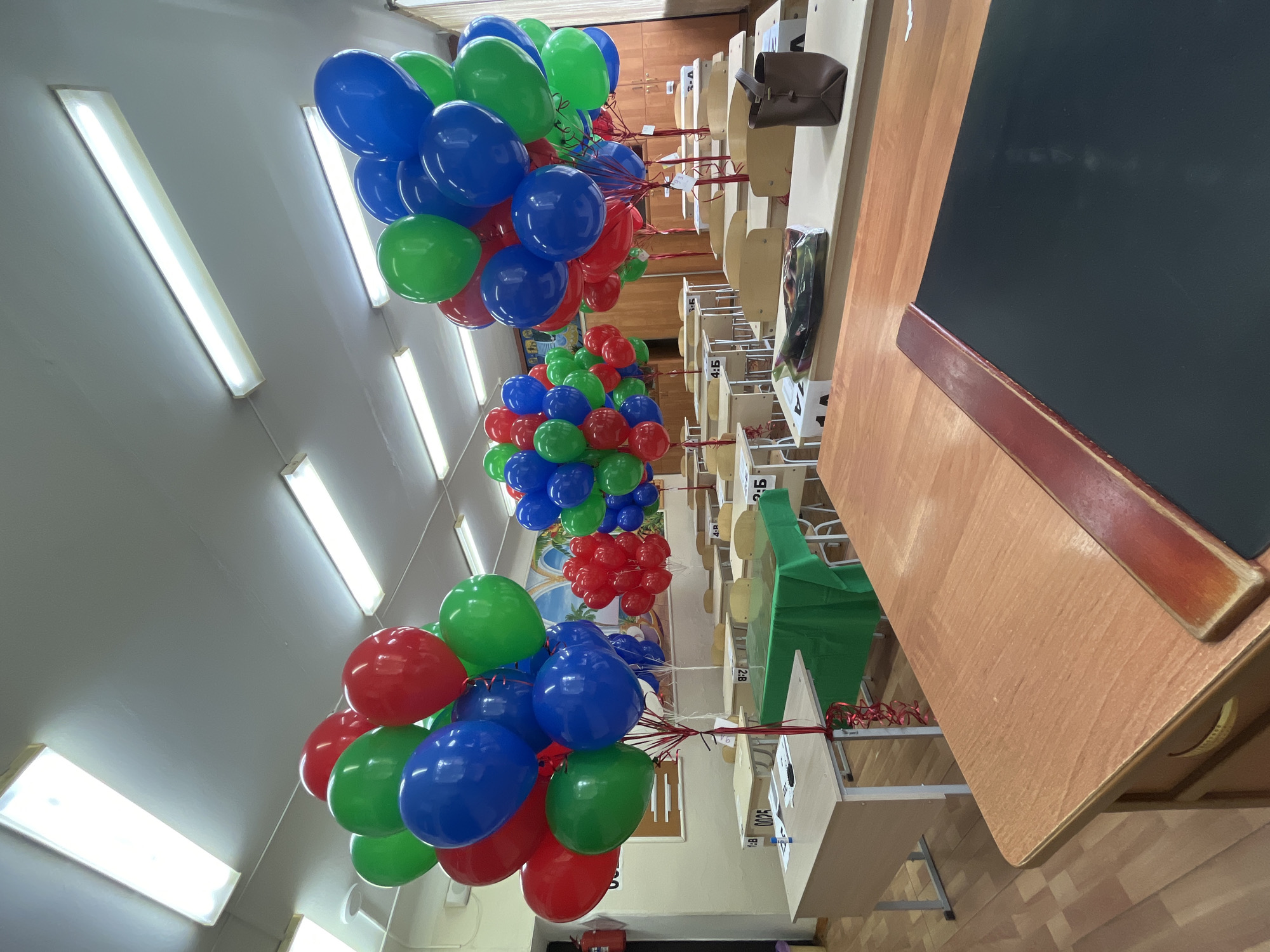 The image shows a classroom in Kaspiysk, Russia. Bunches of red, green, and blue balloons are tied to the backs of student desks. The classroom is illuminated by fluorescent lights. A teacher's desk with a black chalkboard surface is visible in the foreground.