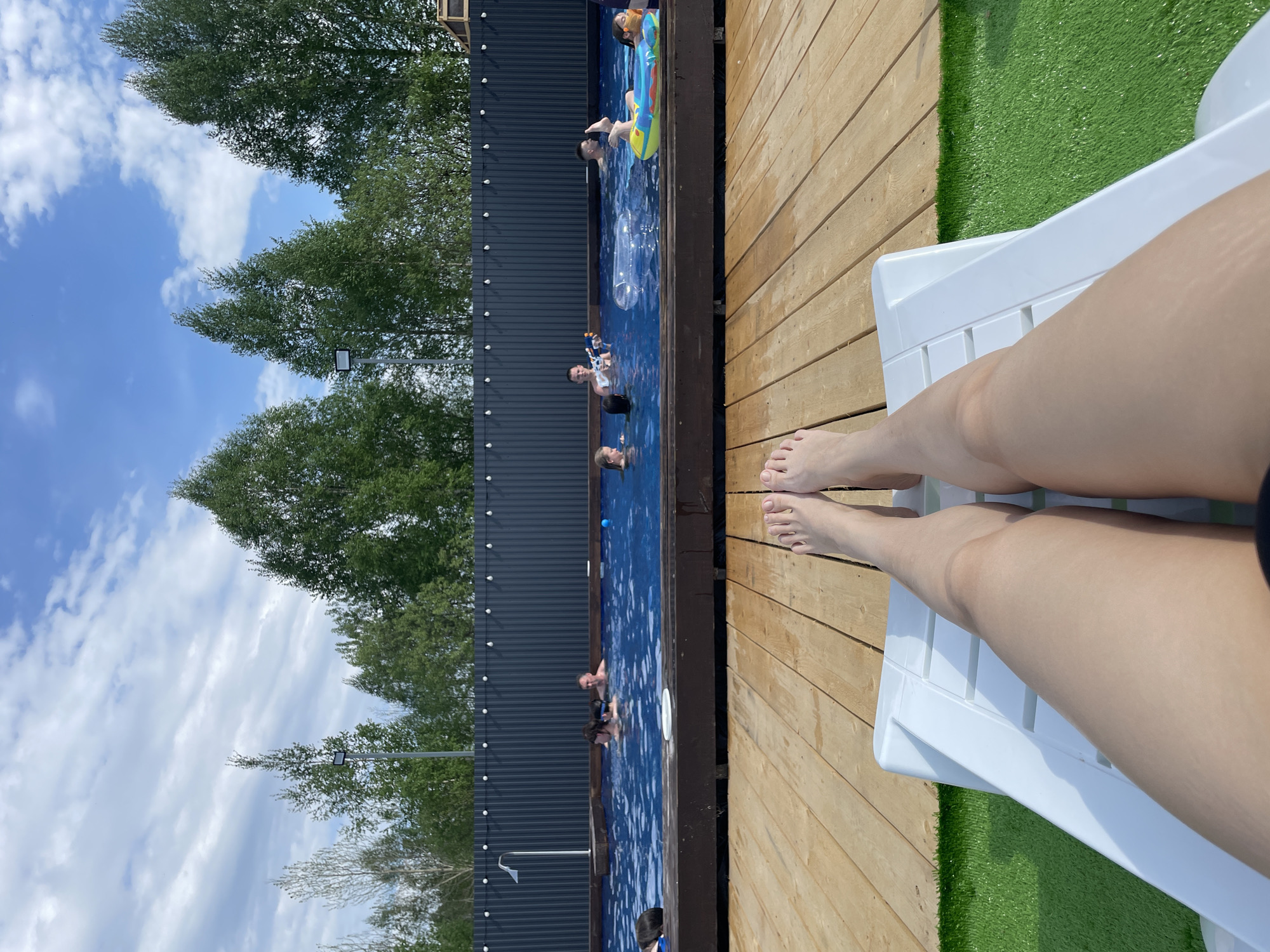 The image shows a person lying on a white plastic lounge chair by a pool. The pool, surrounded by a wooden deck and artificial grass, has several people swimming and floating in it. A dark grey corrugated metal fence borders the pool area, behind which are green trees and a blue sky with clouds. The image is oriented with the sky on the left and the person's legs on the right. The location is Люкшудья, Russia, as stated in the prompt.