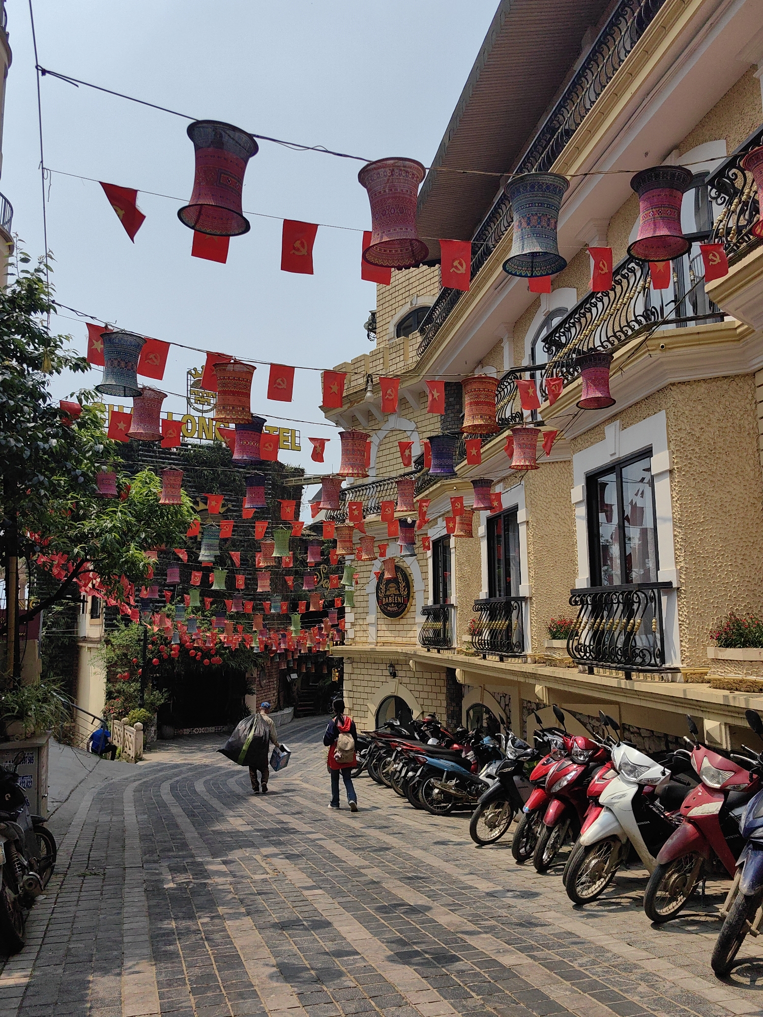 The image displays a decorated street scene in Sa Pa, Vietnam, under a bright, clear sky. The street, paved with dark and light gray interlocking bricks arranged in a wavy pattern, curves slightly to the left in the distance.
On the right, a multi-story building features a textured, light yellow facade with white trim around arched windows and balconies. Each balcony has ornate black wrought-iron railings. A circular sign near the bottom right reads "BABUINI HOTEL." Potted red flowering plants are visible on the balconies.
Overhead, numerous wires crisscross the street, suspending decorative elements. These include strings of small, rectangular red flags, some featuring a yellow star (Vietnamese flag) and others a yellow hammer and sickle (communist symbol). Interspersed among the flags are various cylindrical lanterns in hues of red, blue, purple, and green, displaying intricate patterns. Further down the street, on the left, a building largely obscured by lush greenery bears a prominent yellow sign indicating "HALONG HOTEL."
A line of motorbikes and scooters, predominantly Honda models in colors such as red, white, black, and blue, are parked along the right sidewalk.
Three individuals are present. Two adults are walking away from the viewer down the street. The lead individual carries a large, dark green or black bag over one shoulder and a white box in the other hand. The second individual, following behind, wears a red top, blue jeans, and a light-colored backpack. A third individual, dressed in a blue shirt, is seated or crouching near a low wall and greenery on the left side of the street, further in the background.