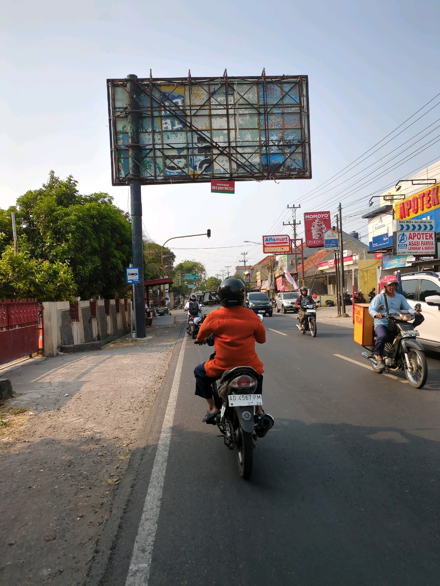 The image shows a street scene in Desa Ngesrep, Indonesia. The viewpoint is from a motorcycle, looking forward at the road and surrounding environment. In the foreground, the rear of a motorcycle with license plate AD 4567 PM is visible, with a person wearing an orange shirt and dark shorts riding it. Further ahead on the same road, several other motorcycles and cars are moving. On the right side of the road, there are shops with signage including "APOTEK" (pharmacy) and "Alfamart". A large, weathered billboard stands on a pole to the left of the road. Trees and utility poles are also present. The sky is clear and blue, suggesting daytime.
