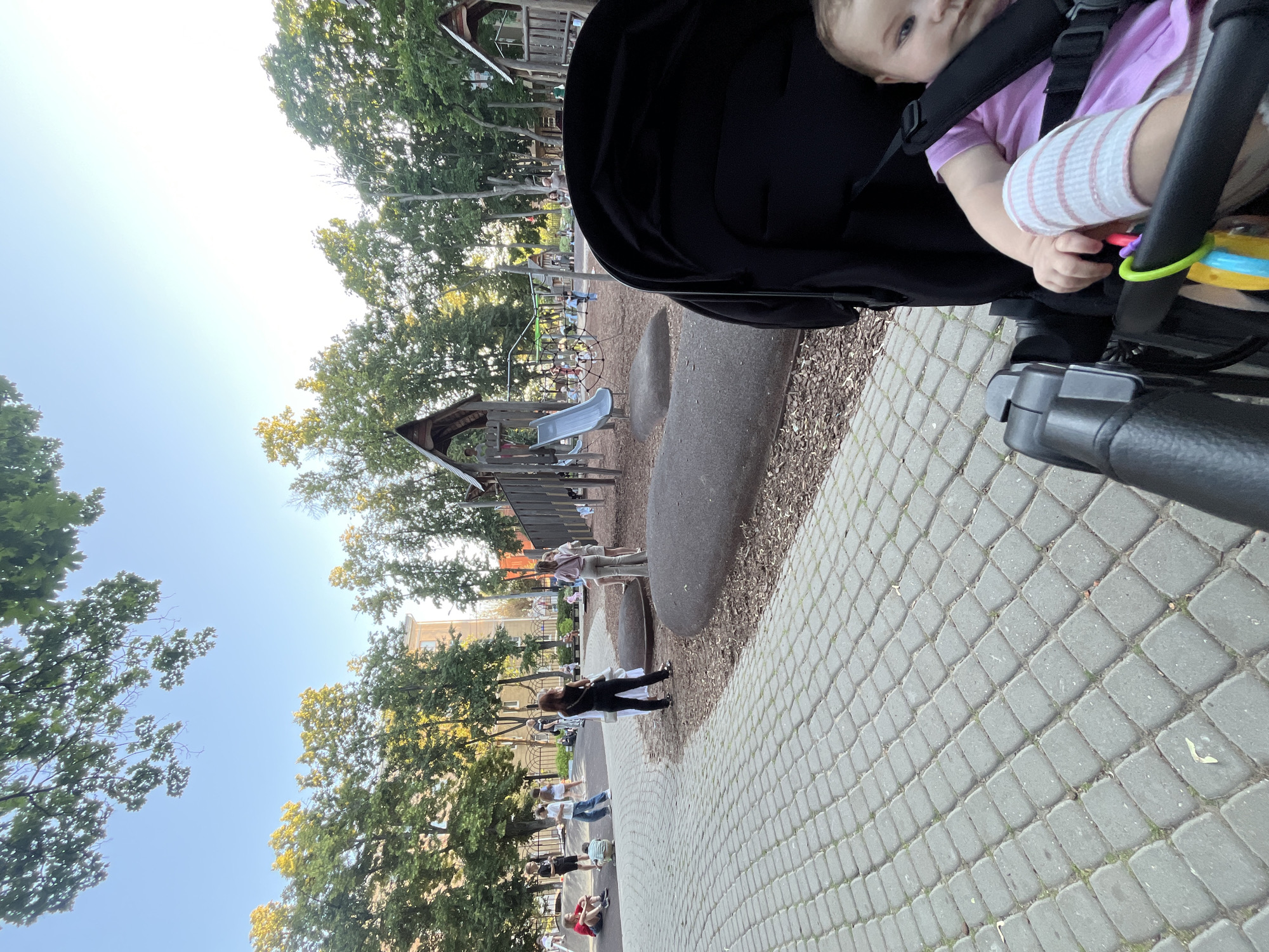 The image depicts a bright, sunny day at a playground in Novaya Usman', Russia. In the foreground, a baby is visible in a stroller, looking towards the camera. The baby is wearing a pink shirt and striped sleeves and has a toy attached to the stroller. Behind the stroller, a paved walkway made of interlocking grey stones stretches into the distance. The playground features a wooden structure with a slide, surrounded by trees and wood chips. Several people are scattered throughout the playground, some standing, some sitting on the ground, and a few appear to be engaged in activities or conversing. The time of day appears to be late afternoon or early evening, judging by the angle of the sun and the lighting. The overall mood seems relaxed and casual, suggesting a typical day at a community park. There is no visible text in the image.