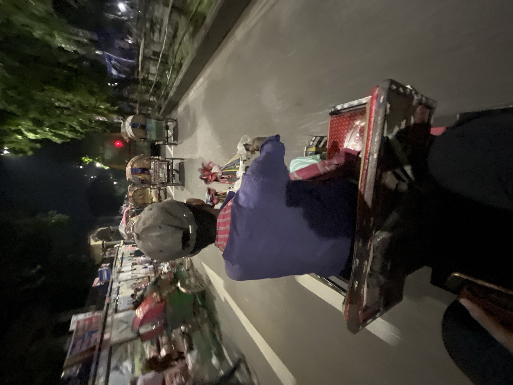 The image depicts a scene from the city of Dhaka, Bangladesh, taken from a high angle, possibly from a rickshaw or building. The main subject is a person wearing a gray cap and a dark top, positioned in the foreground. The background features a street lined with street vendors displaying various goods, including clothes, toys, and other merchandise. Some people are visible on the street, likely shoppers or passersby. The overall environment suggests a crowded urban area, typical of Dhaka. The lighting indicates it might be evening or late afternoon.