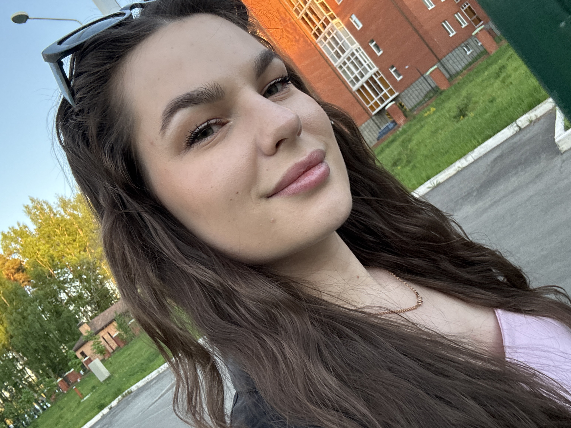 A selfie of a young woman with long brown hair is taken outdoors. She has sunglasses perched on top of her head and is wearing a light pink top with a delicate gold necklace. The background shows a brick building with white-framed windows, a grassy lawn, a paved area, and trees in the distance. The location is likely in a residential area within the city of Chaikovsky, Russia.