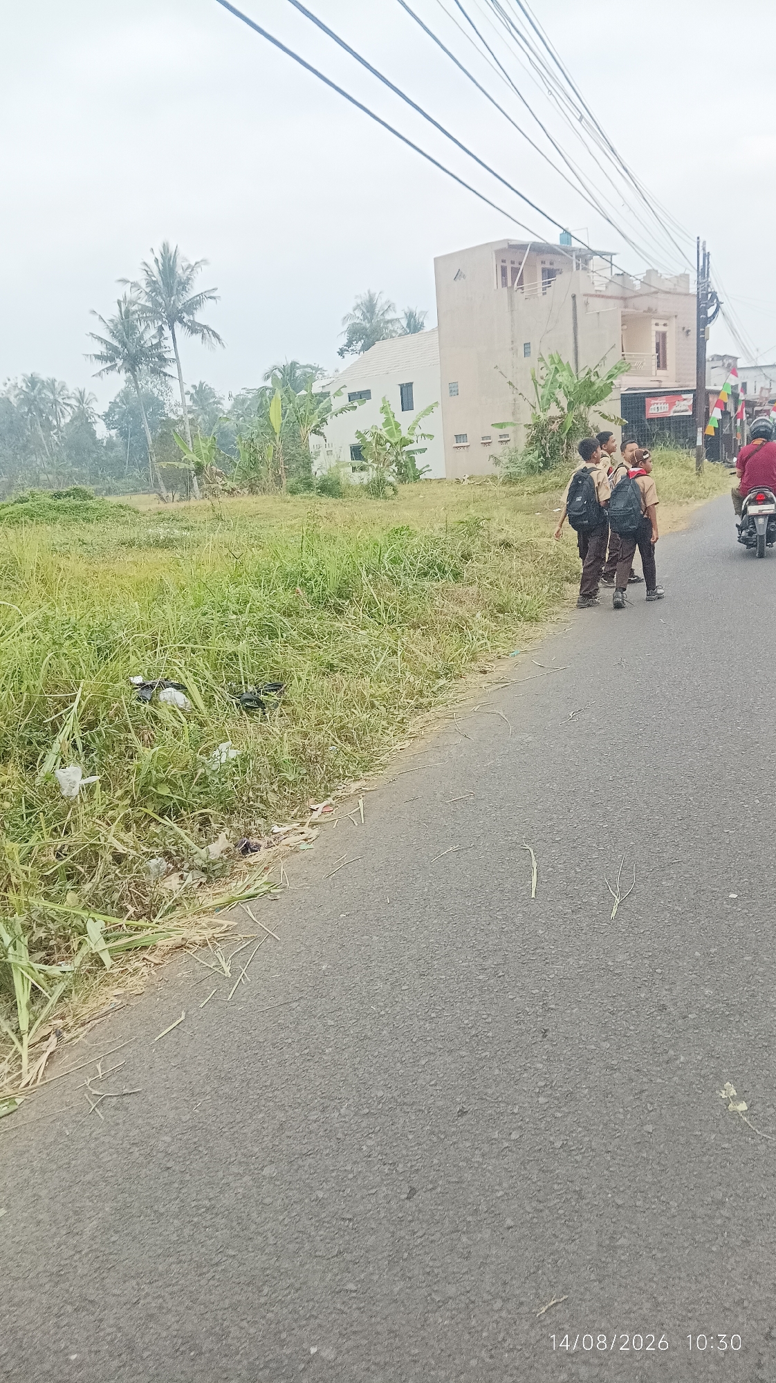 The image depicts a street scene in Tasikmalaya, Indonesia, with a cloudy sky. In the foreground, an asphalt road runs diagonally across the frame. To the left of the road is a grassy field with some scattered trash. In the middle ground, three school children wearing uniforms and backpacks are standing on the side of the road, appearing to be conversing. Behind them, a multi-story building is visible, with some palm trees and other vegetation surrounding it. Further in the background, more buildings and trees can be seen. A person on a motorcycle is riding away from the camera on the right side of the road. The bottom right corner of the image contains a timestamp: "14/08/2026 10:30".