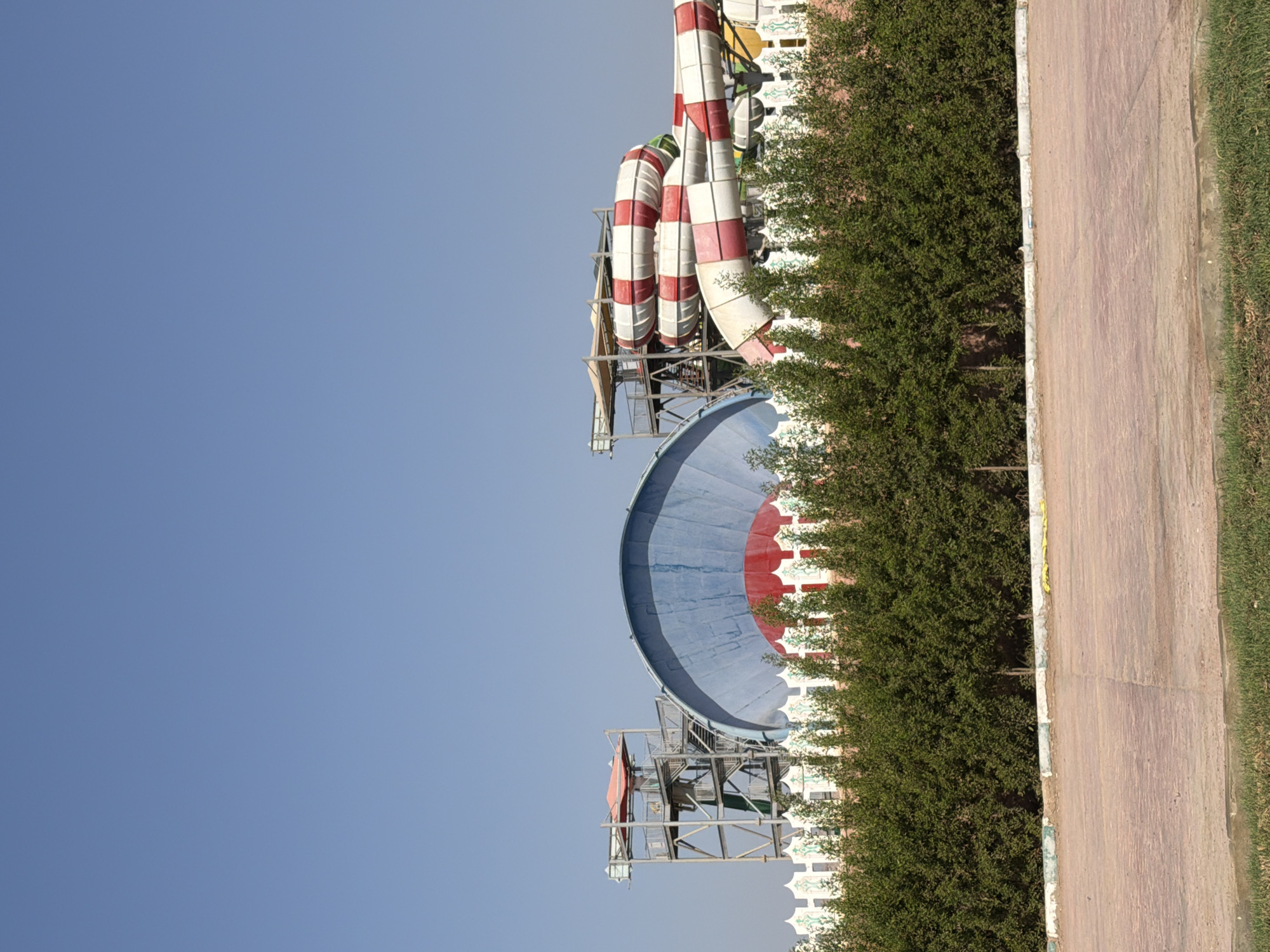 The image depicts a water park in Sharm El Sheikh, Egypt, under a clear blue sky. The foreground features a paved area with a strip of grass. Behind this, a dense line of green bushes forms a barrier. Beyond the bushes, a decorative white fence with green and red accents separates the park's attractions from the viewer. Dominating the background are several large water slides. To the left, a tall metal tower with stairs and a red-roofed platform stands beside a massive, curved blue structure with a red base, resembling a giant fan or dome. To the right, several sections of a red and white checkered water slide twist and turn, with one section extending towards the foreground. The overall scene suggests a sunny day at an amusement park, with no people visible.