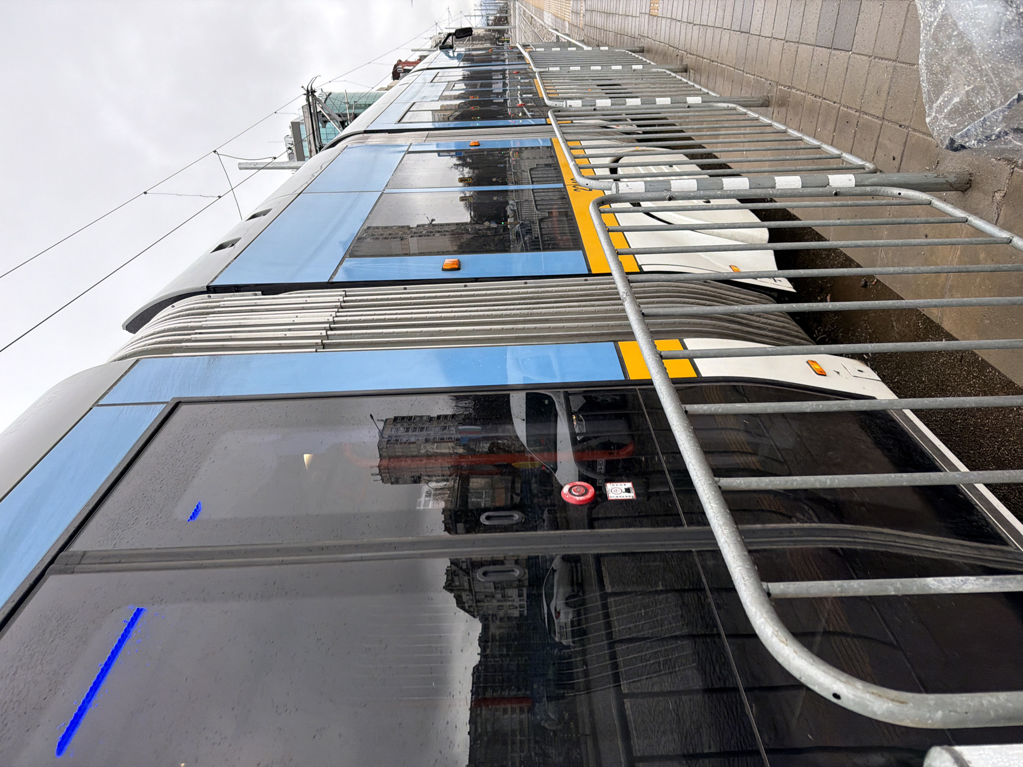 The image depicts a modern, articulated PESA Swing tram, numbered 2010 (or 2040), stationary at an outdoor platform in Sofia, Bulgaria, likely at a street-level stop during overcast weather. The tram, oriented with its left side towards the viewer, is predominantly light blue with a distinct yellow stripe along its lower body and a white undercarriage. Its dark, reflective windows are covered in water droplets, indicating recent or ongoing precipitation.
The windows reflect an urban streetscape, including older, multi-story concrete buildings, some appearing worn, alongside glimpses of newer glass-fronted structures. Reflected vehicles include a white SUV and a white van. A red circular safety light, likely belonging to the tram's exterior, is visible in reflection. Interior blue LED strip lighting is faintly visible through the tram's windows.
In the foreground, a series of galvanized steel crowd control barriers separates the viewer from the tram. These barriers are approximately waist-height, constructed with vertical bars and horizontal rails; some vertical posts are marked with white and black reflective tape. The platform is paved with wet, grey rectangular tiles, featuring a yellow tactile paving strip with raised dots along its edge. Tram tracks are visible between the tram and the platform. Overhead electrical lines for the tram system are present against the light grey, overcast sky. No individuals are clearly visible in the scene.