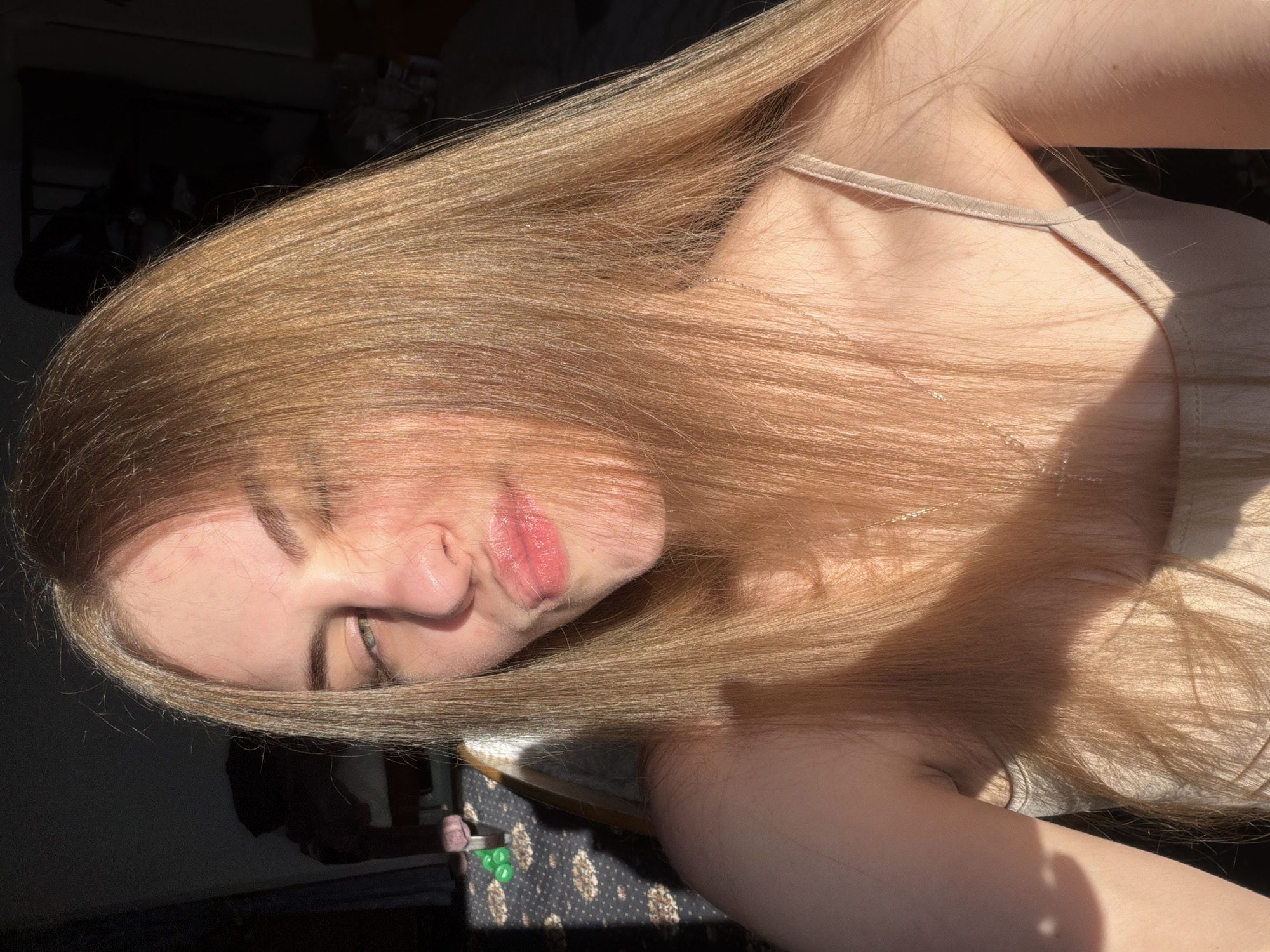 The image is a close-up, indoor selfie of a young woman with long, blonde hair. She is looking towards the camera with one eye partially closed, and her lips are puckered. The lighting suggests it is daytime, with sunlight streaming in from the left, casting shadows across her face and upper body. She is wearing a light-colored, possibly beige, camisole. The background is dimly lit and appears to be a room, with some objects visible but out of focus. There is no information in the image to determine the location in the city of Dnipro, Ukraine.