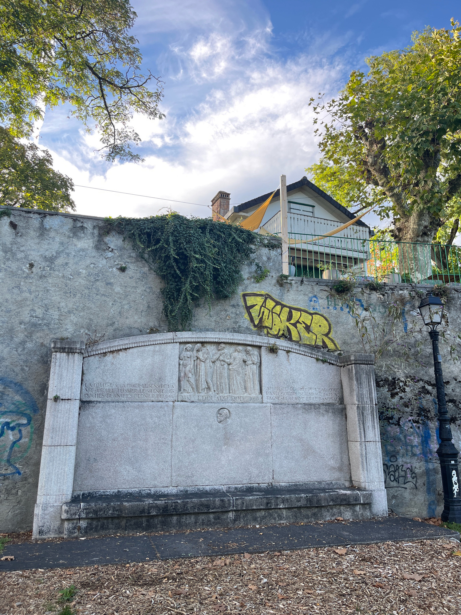 The image captures a section of a stone monument and retaining wall in Nyon, Switzerland. The monument is intricately carved with bas-relief figures of people and text. Graffiti, including the word "MAVER" in yellow spray paint, is visible on the stone wall above and to the right of the monument. Green ivy grows on the wall. A black lamppost stands to the right of the monument. In the background, a white building with a balcony and a sloped roof is partially visible, along with trees and a cloudy sky. The ground in front of the monument is paved and covered with fallen leaves.