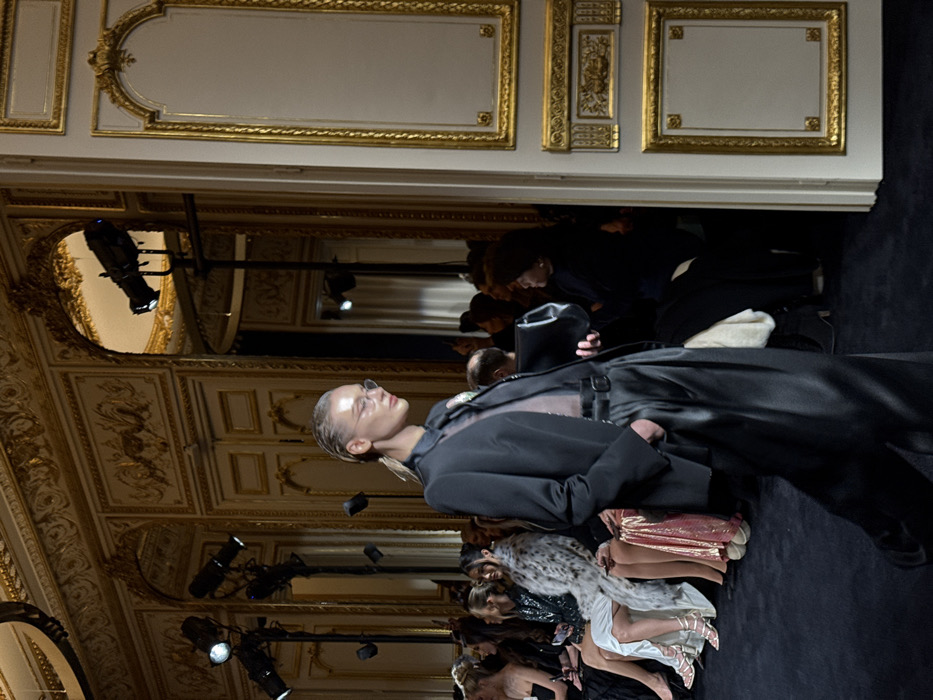 A fashion show setting features a model in a dark, belted outfit walking past the front row. The ornate room has gilded molding and lighting fixtures. Attendees are seated, dressed in a variety of styles and colors. The scene is captured at a unique angle, focusing on the model and front row while the room's architecture fills the background.