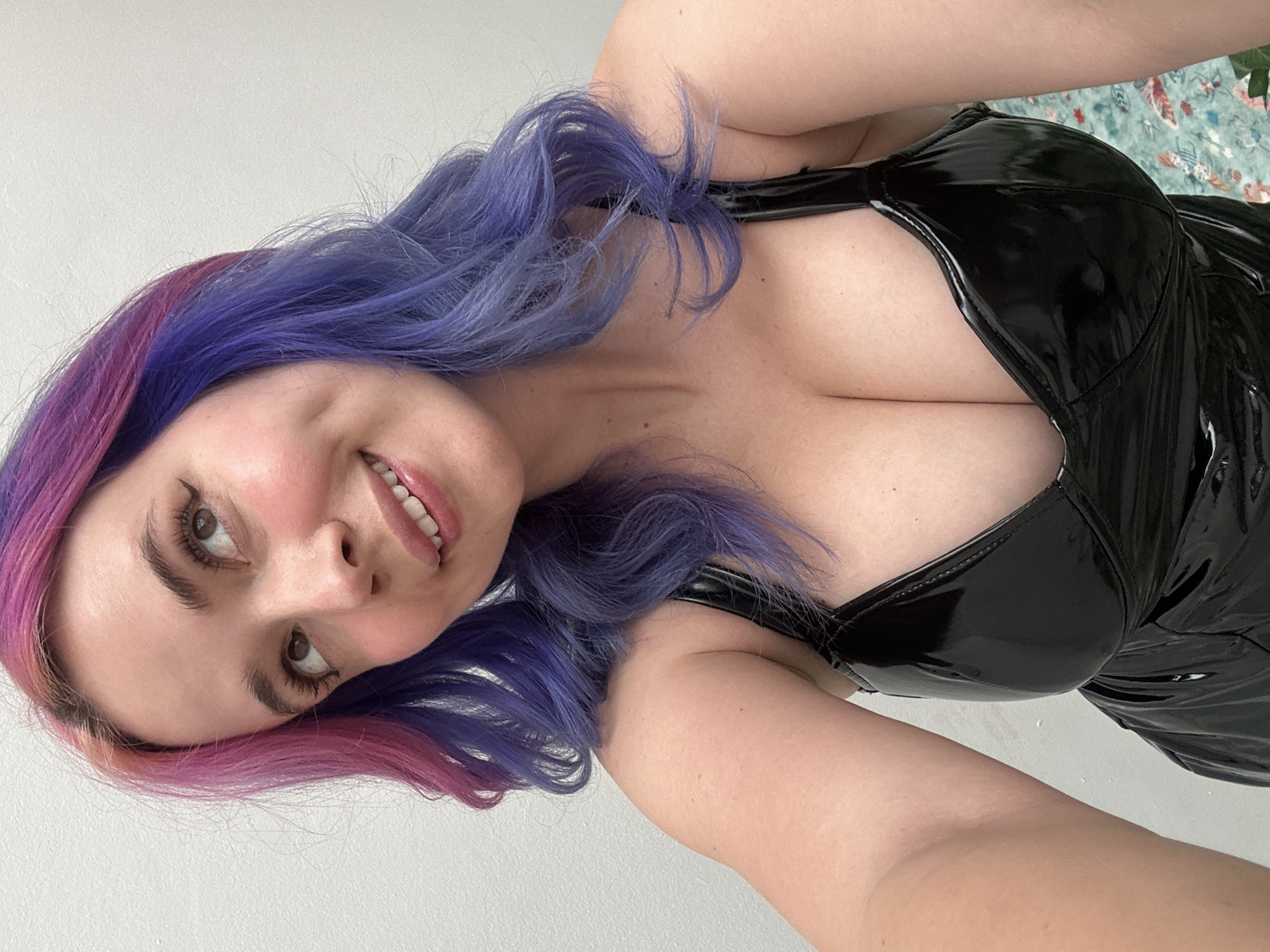 A close-up, rotated selfie shows a young woman smiling and posing indoors. Her head is tilted to her left, and she looks slightly upwards and to the right of the camera. She has fair skin, prominent rosy cheeks, and brown eyes. Her long, wavy hair is dyed in vibrant shades, transitioning from purple on one side to pink or magenta on the other. She has well-defined eyebrows and appears to be wearing eyeliner and mascara. Her lips are slightly parted in a smile, revealing white teeth.
The woman is wearing a fitted, black, shiny top, likely made of latex or PVC, featuring a low-cut neckline that exposes her décolletage. The glossy material reflects light, creating highlights. Her bare arm is visible in the foreground, extending from the bottom right.
The background is predominantly a plain, light-colored wall, suggesting an indoor setting like a room in a home. A small section of a blurry, light green surface with a subtle floral or abstract pattern is visible in the upper right corner, possibly a blanket or cushion. The lighting is bright and even, indicating daytime or well-lit conditions. The woman's expression conveys happiness and confidence. There is no visible text in the image. While the location context is given as Zagreb, Croatia, the image itself does not contain any specific visual cues to identify this city.