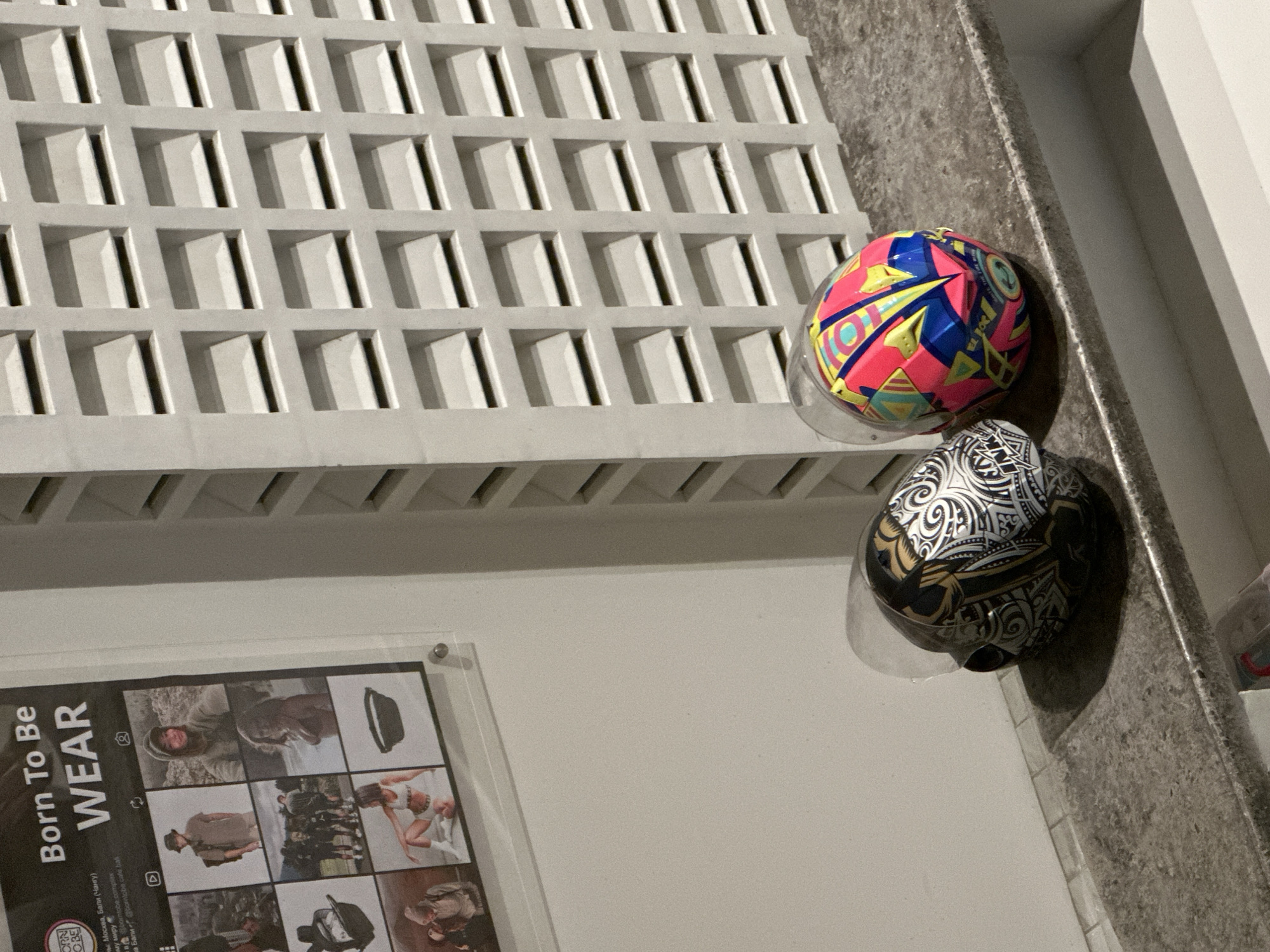 The image shows two helmet-like objects with transparent visors resting on a countertop in front of a patterned wall. The top object is a multi-colored sphere, primarily pink, blue, yellow, and green. Below it, the second object has a black and white pattern. A framed poster with the words "Born To Be WEAR" is visible on the wall to the left of the objects. The poster mentions "Москва, Бали (Чангу)" along with Instagram handles related to "borntobe" and refers to the city of Mengwi, Indonesia. A window is visible on the right side of the image.