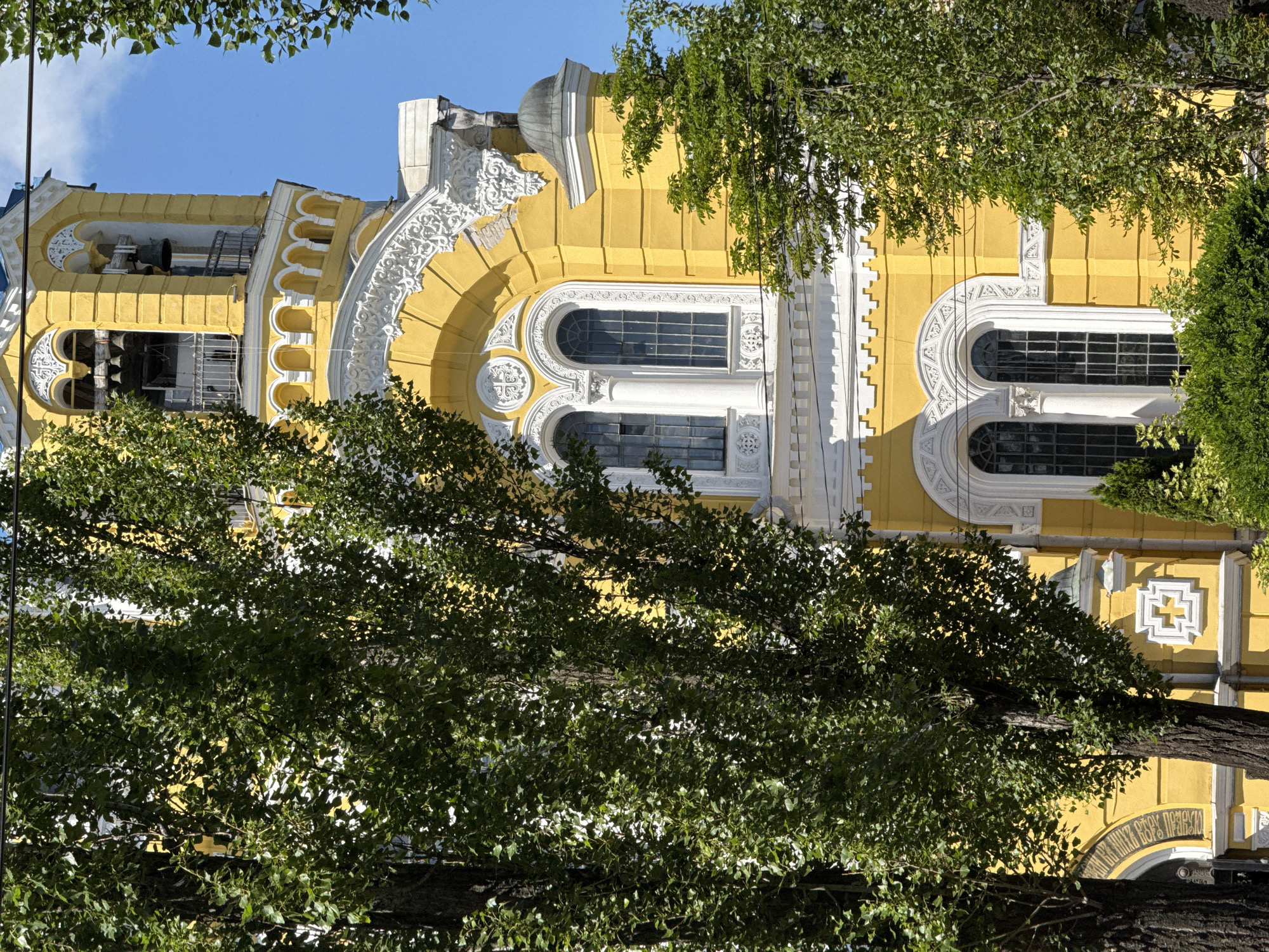 This is a medium-shot, eye-level, vertical photograph of the exterior of St. Michael's Golden-Domed Monastery in Kyiv, Ukraine. The main subject is a section of the monastery's yellow facade, featuring white architectural details and arched windows. The top of the bell tower is visible, with bells hanging in its openings. The scene is framed by the dense green foliage of two large trees in the foreground and on the right side of the frame, partially obscuring the building. The sky above is a clear, bright blue. The sunlight casts shadows from the trees onto the building, highlighting its textures and architectural features. There are no people or animals present in the frame.
