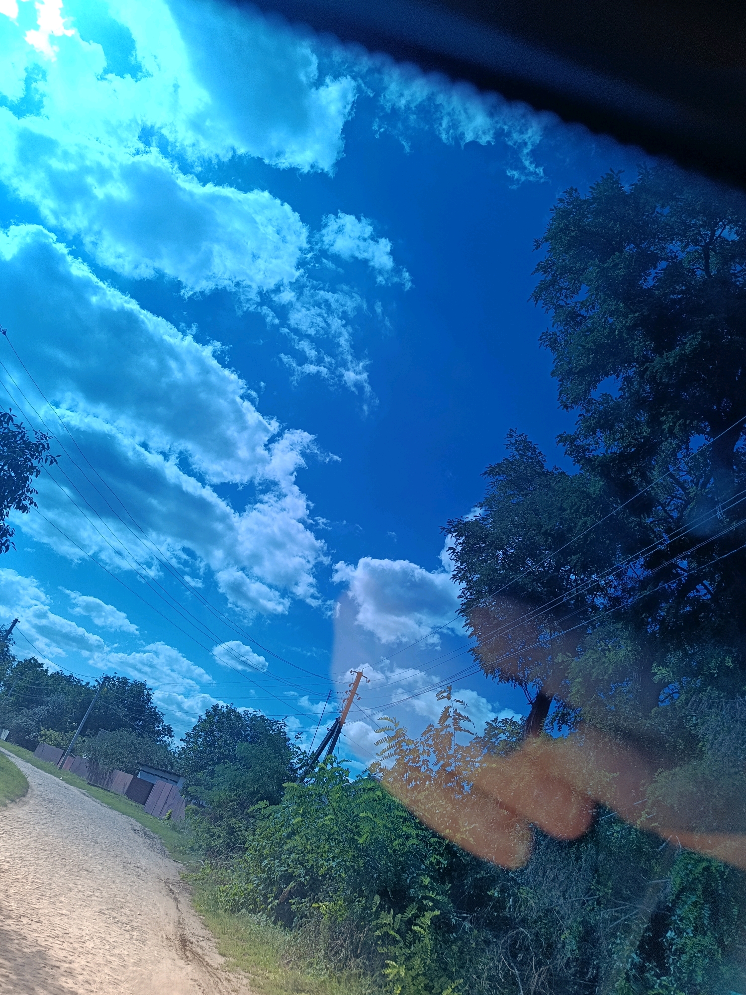 The image depicts a dirt road winding through a rural area, likely in Popivka, Ukraine, under a bright blue sky filled with scattered white clouds. Dense green foliage and trees line the right side of the road, with a few buildings and a utility pole visible in the background. The perspective suggests the photo was taken from a vehicle moving along the road. The overall scene conveys a sense of everyday life in a countryside setting, with natural elements dominating the landscape.