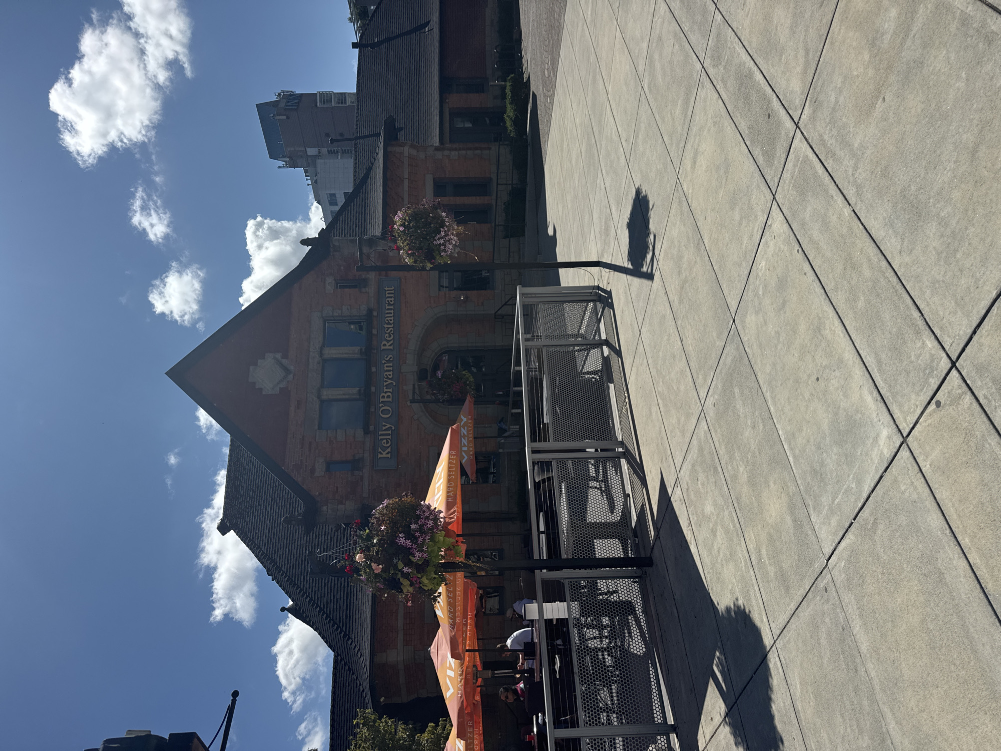 The image shows the exterior of Kelly O'Bryan's Restaurant in New Westminster, Canada. The building is made of red brick with a dark-tiled roof and a prominent gable. In front of the restaurant, there is an outdoor seating area with orange umbrellas and metal tables and chairs. Several people are seated at the tables, some appearing to be dining. Hanging flower baskets are visible, adding a decorative element. The ground in the foreground is paved with concrete tiles. In the background, other buildings, including a tall modern structure, are visible under a bright blue sky with scattered clouds. The scene suggests a pleasant, sunny day with outdoor dining in progress.