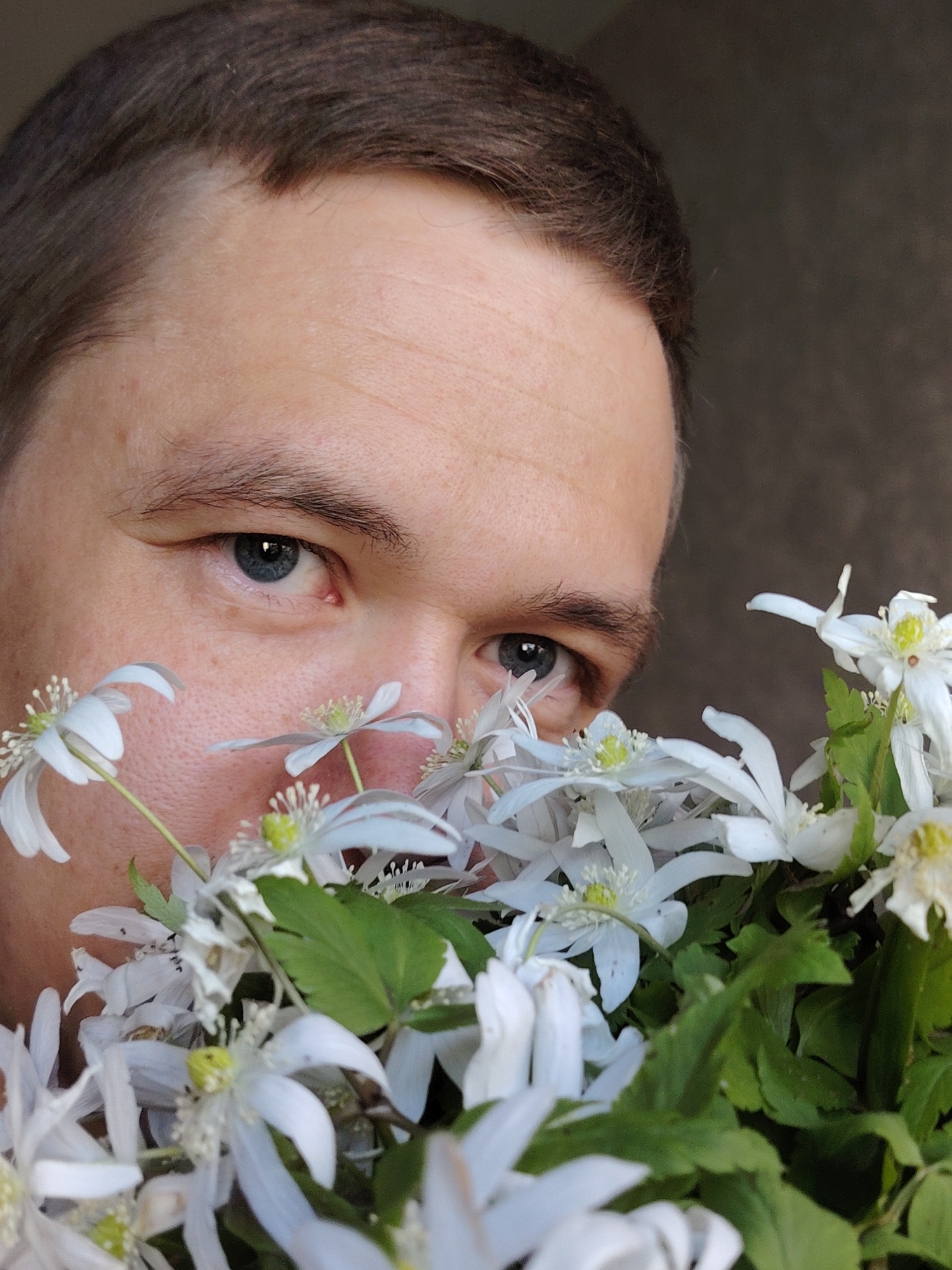 A close-up image features a person with light skin, partially obscured by a dense bouquet of white flowers. The upper part of the person's face is visible, including a forehead with dark brown hair and piercing blue eyes that look directly at the viewer. The eyes convey a gentle, observant, and perhaps slightly shy or playful expression.
The flowers are small, white, and delicate, with distinct yellow-green centers and vibrant green leaves. They appear fresh, possibly like spring wildflowers, creating a natural screen in the foreground. Soft, natural light illuminates the scene, suggesting daytime and contributing to a serene atmosphere. The background is dark and blurred, providing a shallow depth of field that keeps focus on the person and flowers. There is no visible text in the image. The scene captures a moment of quiet interaction with nature, likely in Chaikovsky, Russia.
