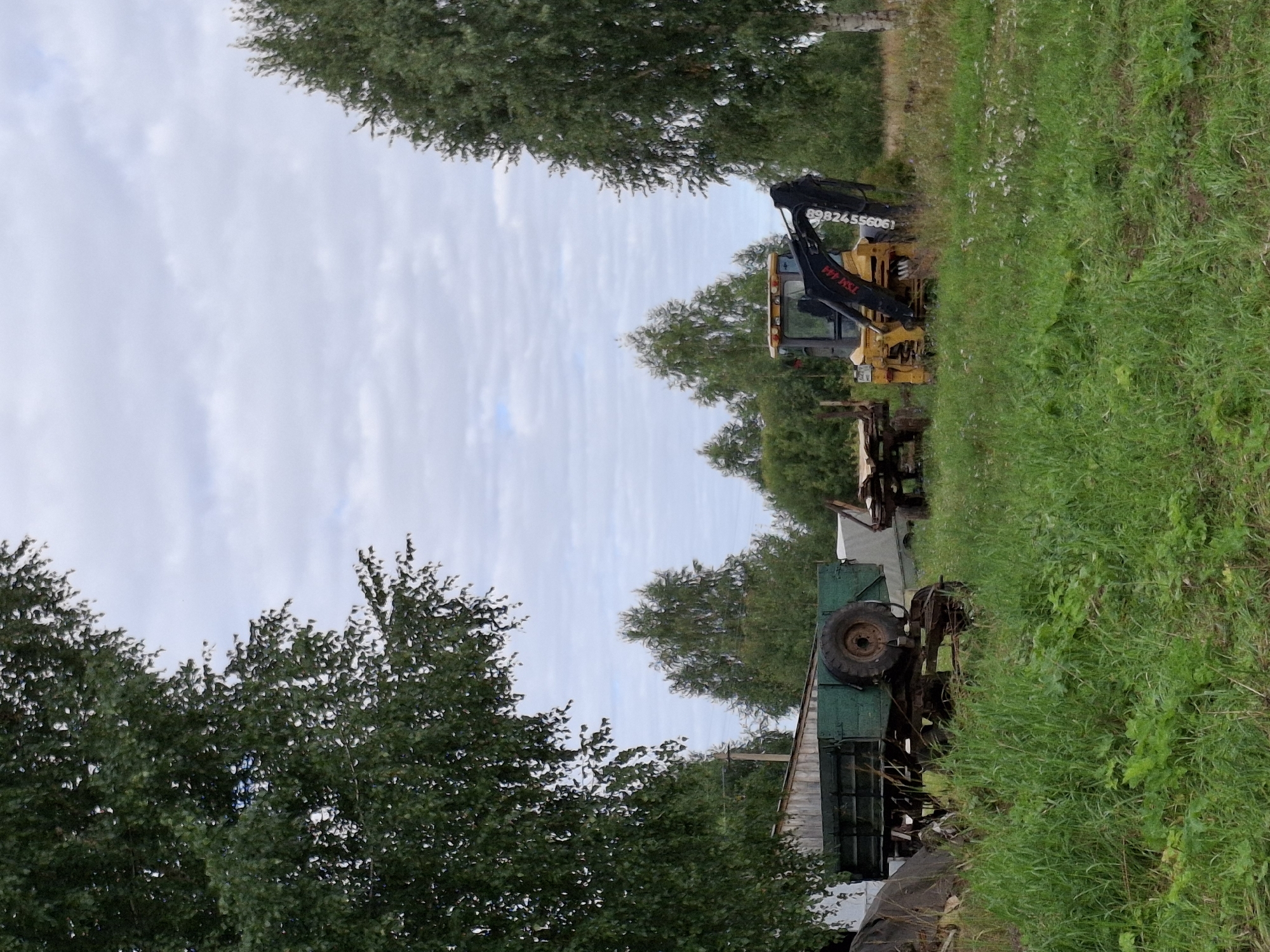 The image depicts an outdoor scene in Plishkino, Russia, featuring agricultural equipment and natural vegetation. In the foreground, tall green grass and various plants cover the ground. To the left, a weathered green trailer with a large tire is parked. Behind it, a section of a wooden structure, possibly a shed or barn, is visible. Further to the right, a yellow backhoe loader is positioned, with its black arm extended. The background consists of a line of trees, with the sky above showing scattered clouds. The overall impression is one of a rural or semi-abandoned area.