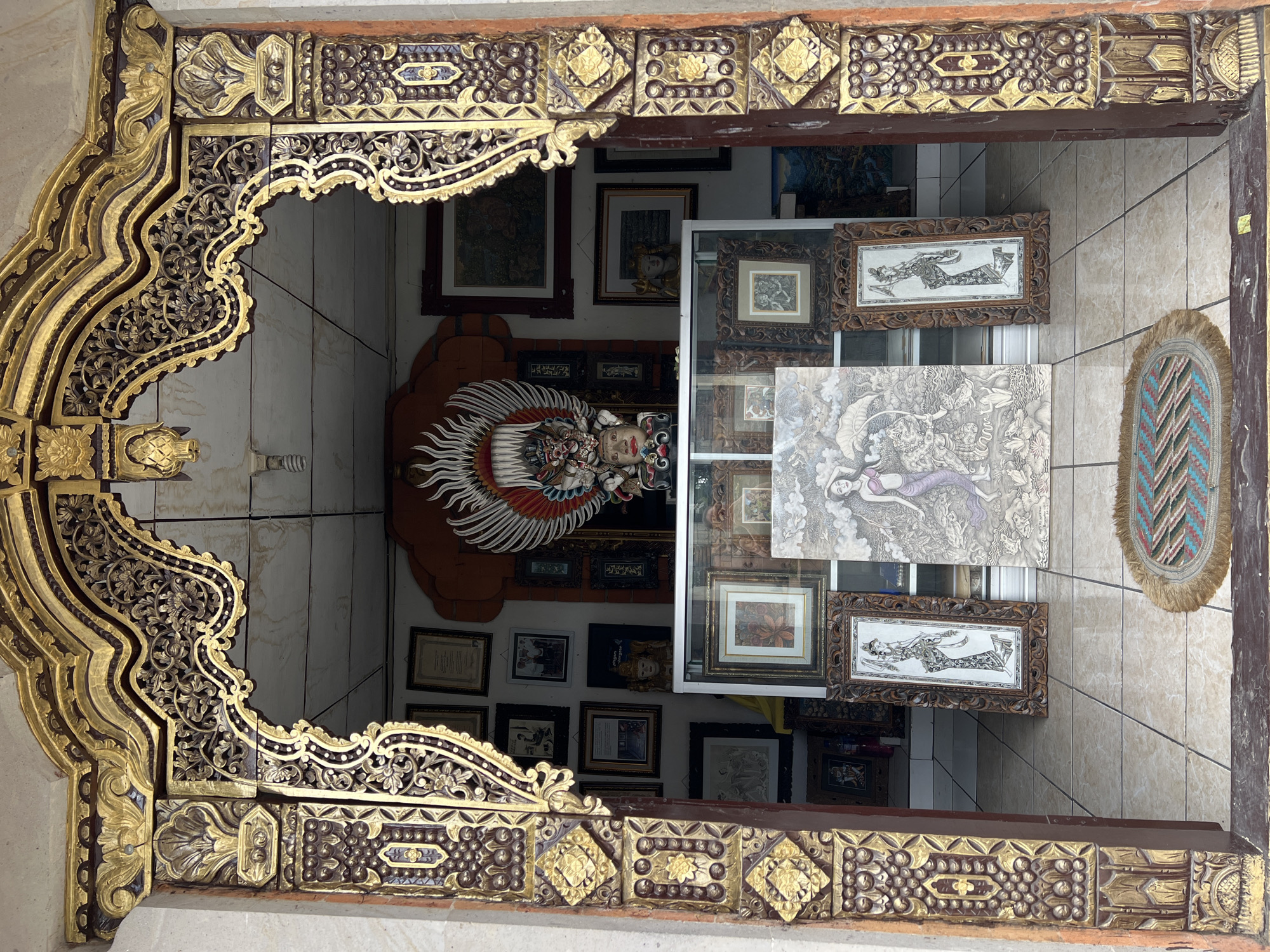 View through an intricately carved, gold-painted and dark-stained wooden frame. The frame likely belongs to a shop or gallery in the Ubud District, Indonesia. Inside the building, various framed artworks are displayed on the walls, including traditional Balinese paintings and carvings. A large Garuda statue with red and white wings dominates the background. The building has tiled floors and walls, giving it a clean, commercial appearance. The scene is viewed from the exterior through an open doorway or window.