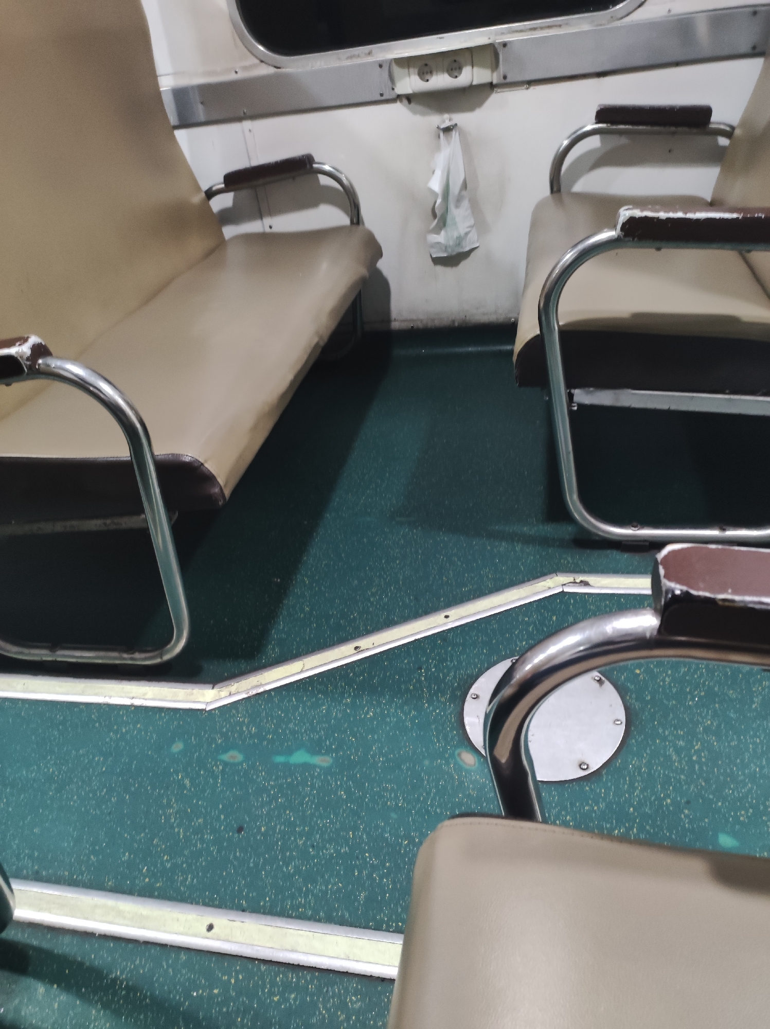The image shows the interior of what appears to be a train or bus, with rows of beige seats and a green speckled floor. The seats have chrome metal frames and armrests with dark brown padding. There is a white cloth hanging from the wall above an electrical outlet. The environment is indoors, with overhead lighting creating shadows. The scene is clean and orderly, suggesting a functional public transportation setting. The location is specified as Talun, Indonesia, however, there is no visual information in the image to confirm or deny this specific location.