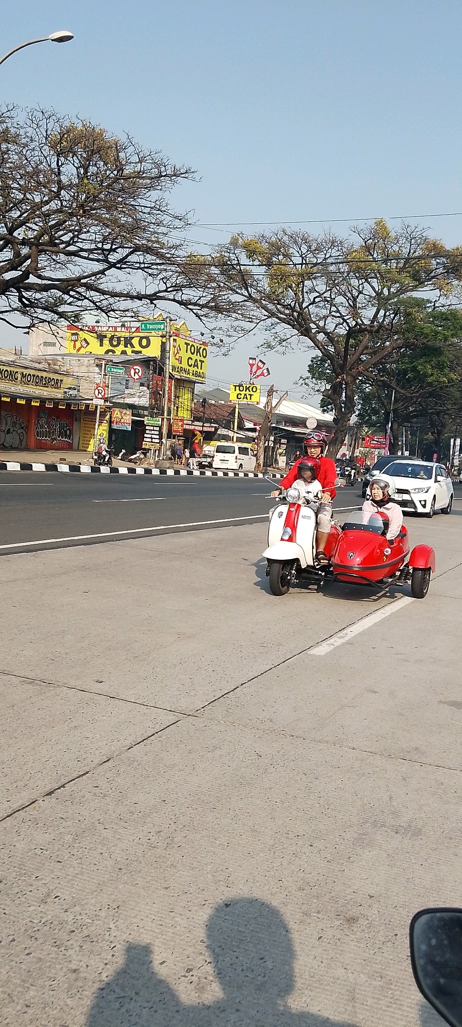 In Gunung Putri, Indonesia, on a clear day, a man and a woman ride a red and white scooter with a red sidecar. The man, wearing a red shirt and helmet, drives the scooter. The woman, in a pink top and helmet, sits in the sidecar. They are on a paved road, with trees and buildings lining the street. In the background, there are several shops with yellow signs, including "TOKO CAT" and "KONG MOTORSPORT." A white car is also visible on the road. The sky is a light blue, and the sun casts shadows on the road.