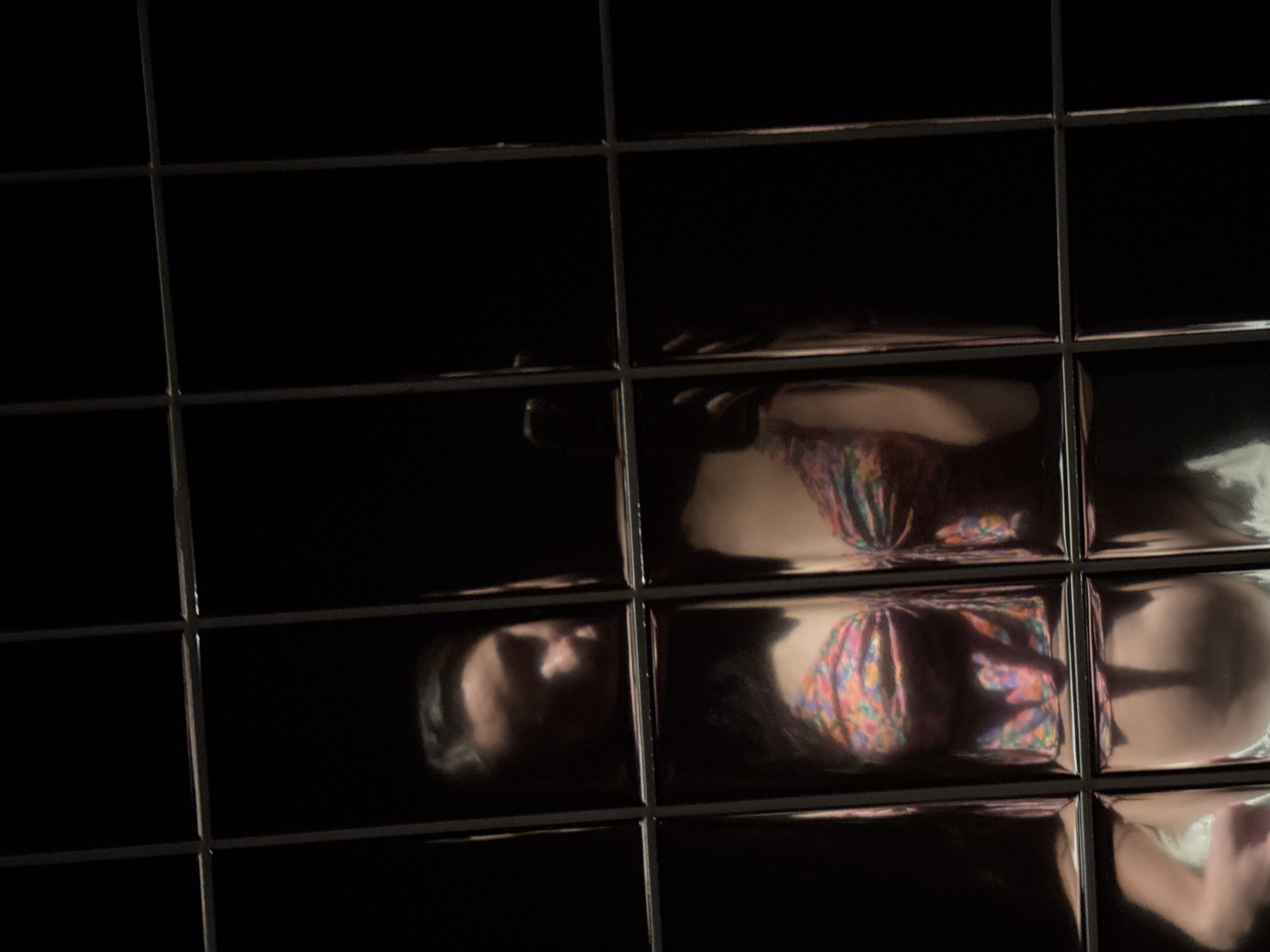 The image shows a reflection of a person in a grid of glossy, dark tiles. The person appears to be lying down and is wearing a patterned top. The reflection is fragmented by the grid lines of the tiles, and some of the tiles are dark and do not reflect the image.
There is no indication of location in Kazan, Russia.
