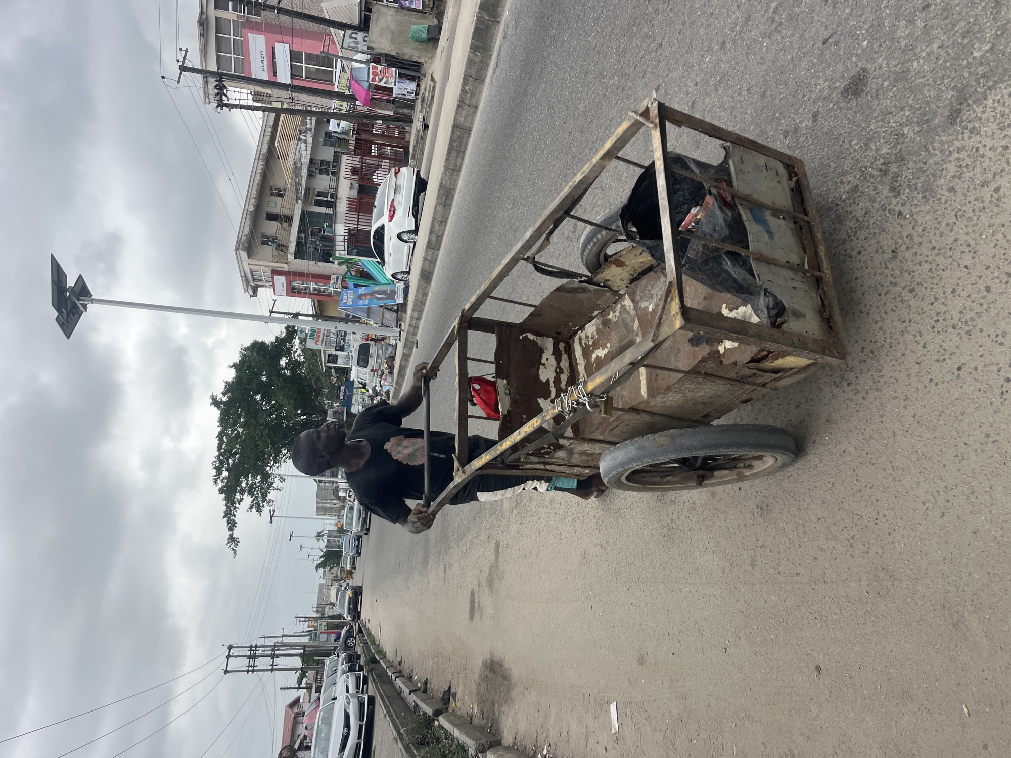 A man is shown from the chest down, pushing a metal cart with two wheels on a paved road. The cart is rusty and appears to be carrying some items, including a black plastic bag. The man is wearing a dark t-shirt and patterned pants. The road is lined with buildings and parked cars. Power lines are visible overhead, and there is a street light. The sky is overcast. The scene is consistent with a street in Ado, Nigeria.
