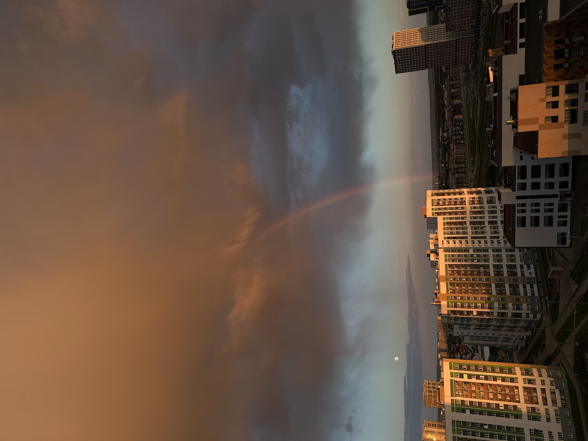 The image depicts an urban landscape in Yekaterinburg, Russia, at twilight. Buildings dominate the foreground, their facades illuminated by the setting sun. A rainbow arcs across the sky, framed by storm clouds tinted in warm hues from the sunset. A moon is also visible in the sky. The backdrop consists of a sprawling cityscape with additional buildings. The overall scene suggests a blend of natural phenomena and urban development.