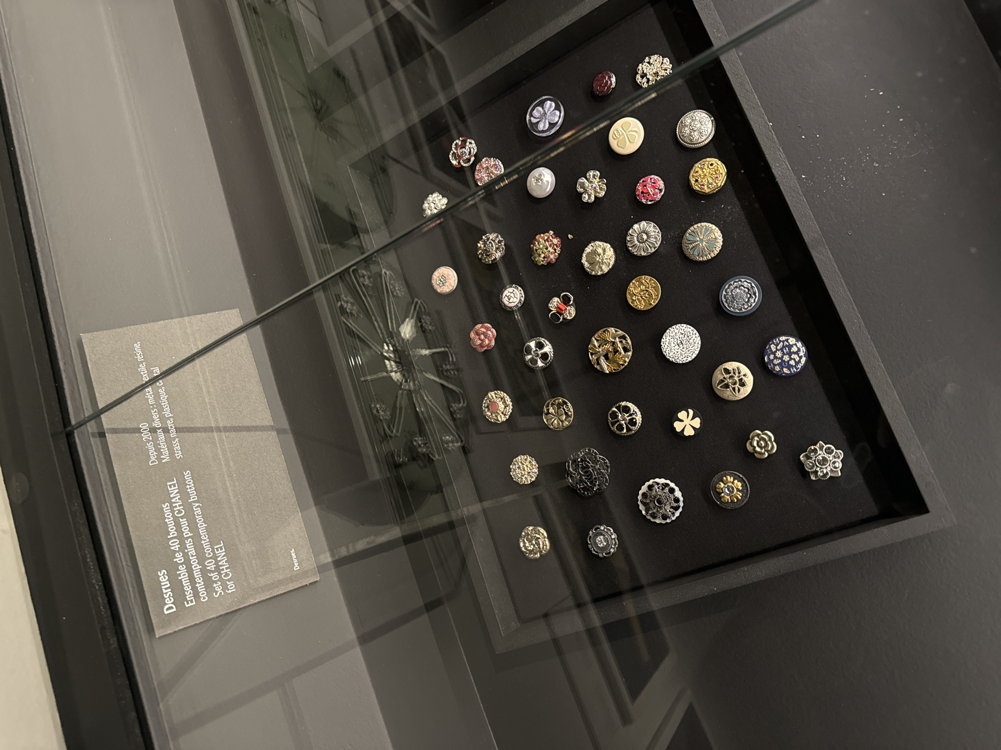 The image shows a framed collection of approximately 40 contemporary Chanel buttons, displayed behind glass, likely in a museum or gallery setting. A label on the display indicates the buttons are a set of 40 contemporary buttons for Chanel by Desrues. The label also mentions the date, since 2000, and various materials: metal textile, resin, strass, nacre, plastic, crystal. The display itself is a black tray set within a darker frame. The lighting is somewhat dim, creating reflections on the glass. The location is likely in Paris, France, given the French language on the label and the historical association of Chanel with Paris.