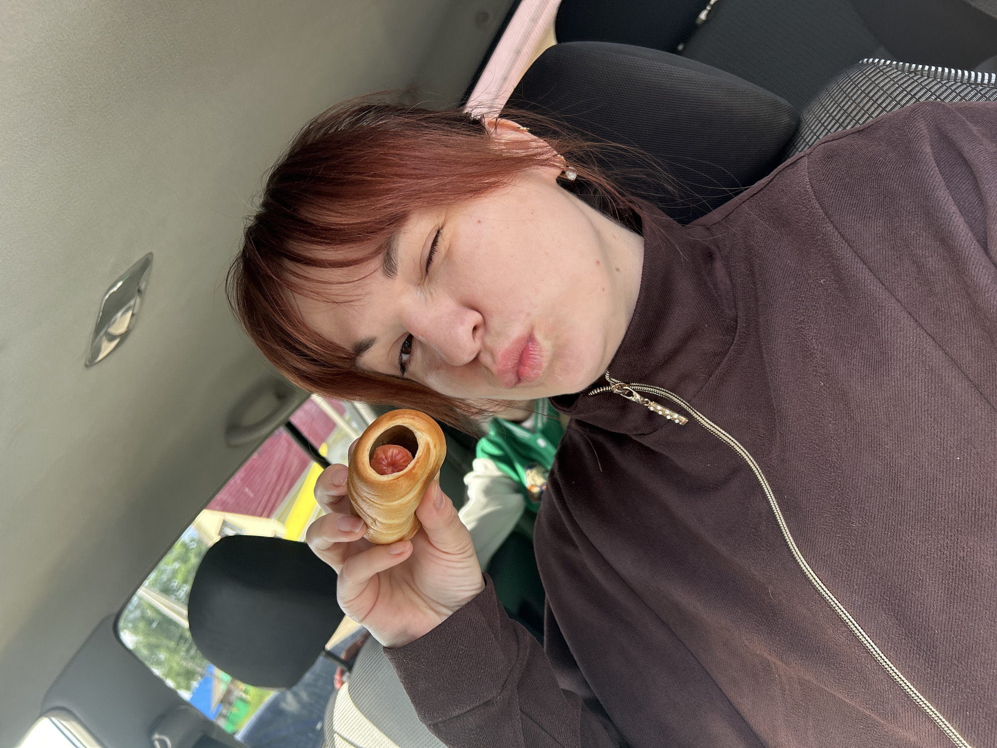 A woman with auburn hair is sitting in a car, holding a sausage roll in her left hand. She is winking and puckering her lips towards the camera. She is wearing a brown, zip-up jacket. Behind her, there are car seats and a glimpse of the outside through the window. Based on the buildings seen through the window, the location is most likely in a residential area, possibly in Фоки, Russia.