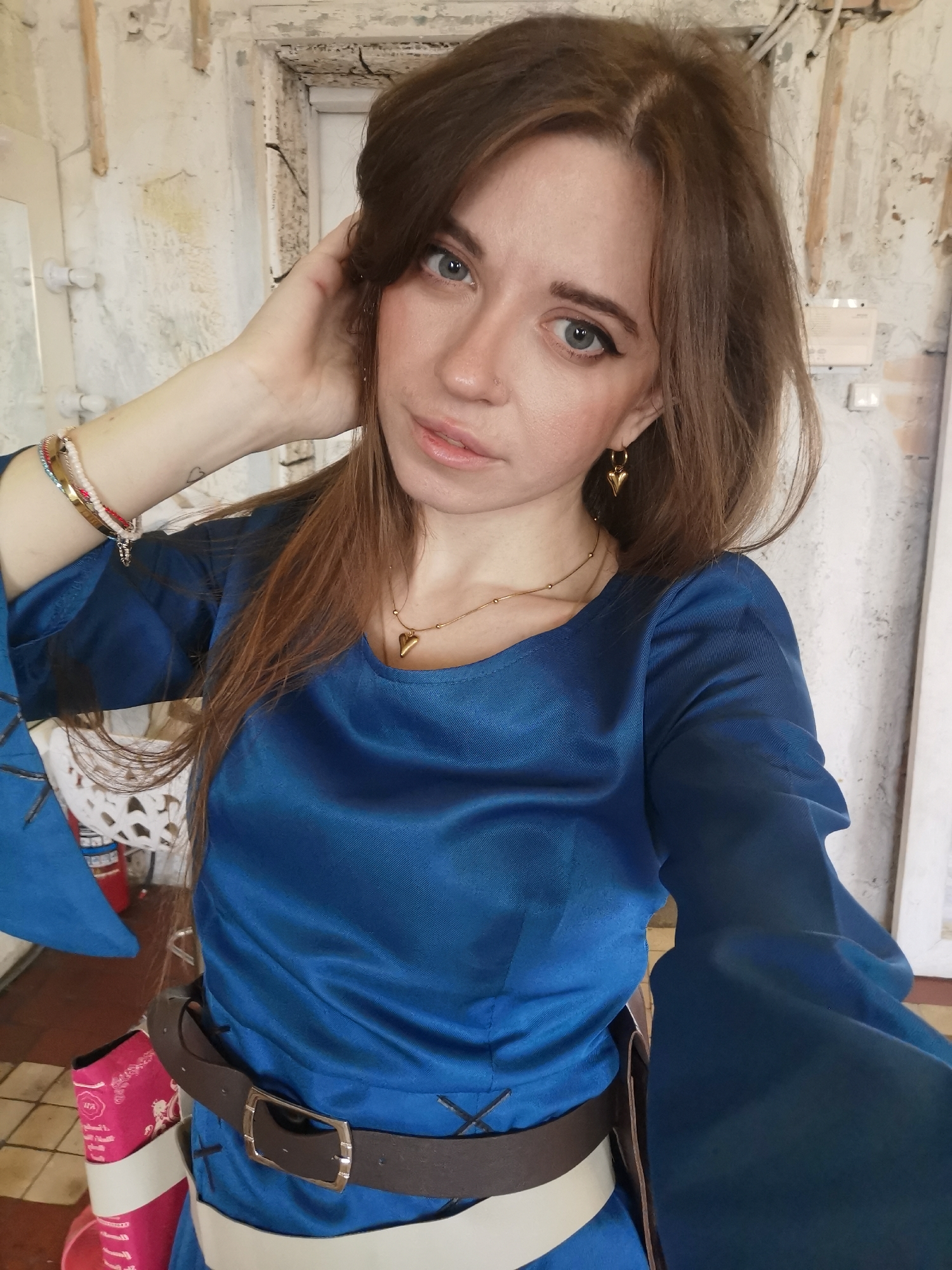 A waist-up shot shows a woman with long brown hair posing indoors. She is wearing a blue, long-sleeved dress with a brown leather belt. She is wearing gold heart-shaped earrings and a necklace with a heart pendant. Her left hand is touching the left side of her head. Her eyes are directed toward the camera, and she has a slight smile. The background appears to be a dressing room or similar space with white, peeling walls and exposed fixtures. The location is Moscow, Russia.